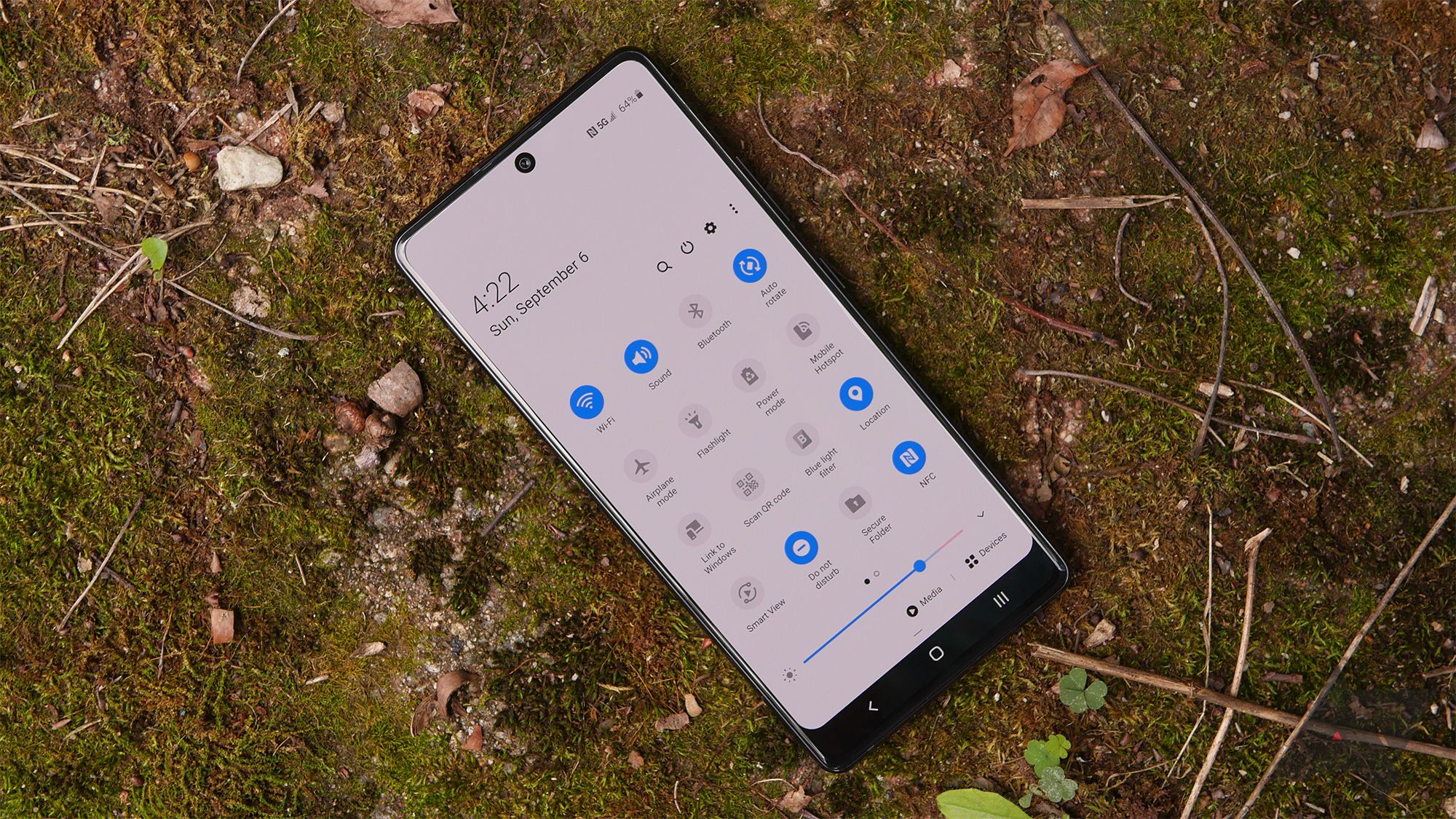 A Samsung Galaxy A71 rests on a mossy stone. The Quick Settings pulldown appears on the display. 