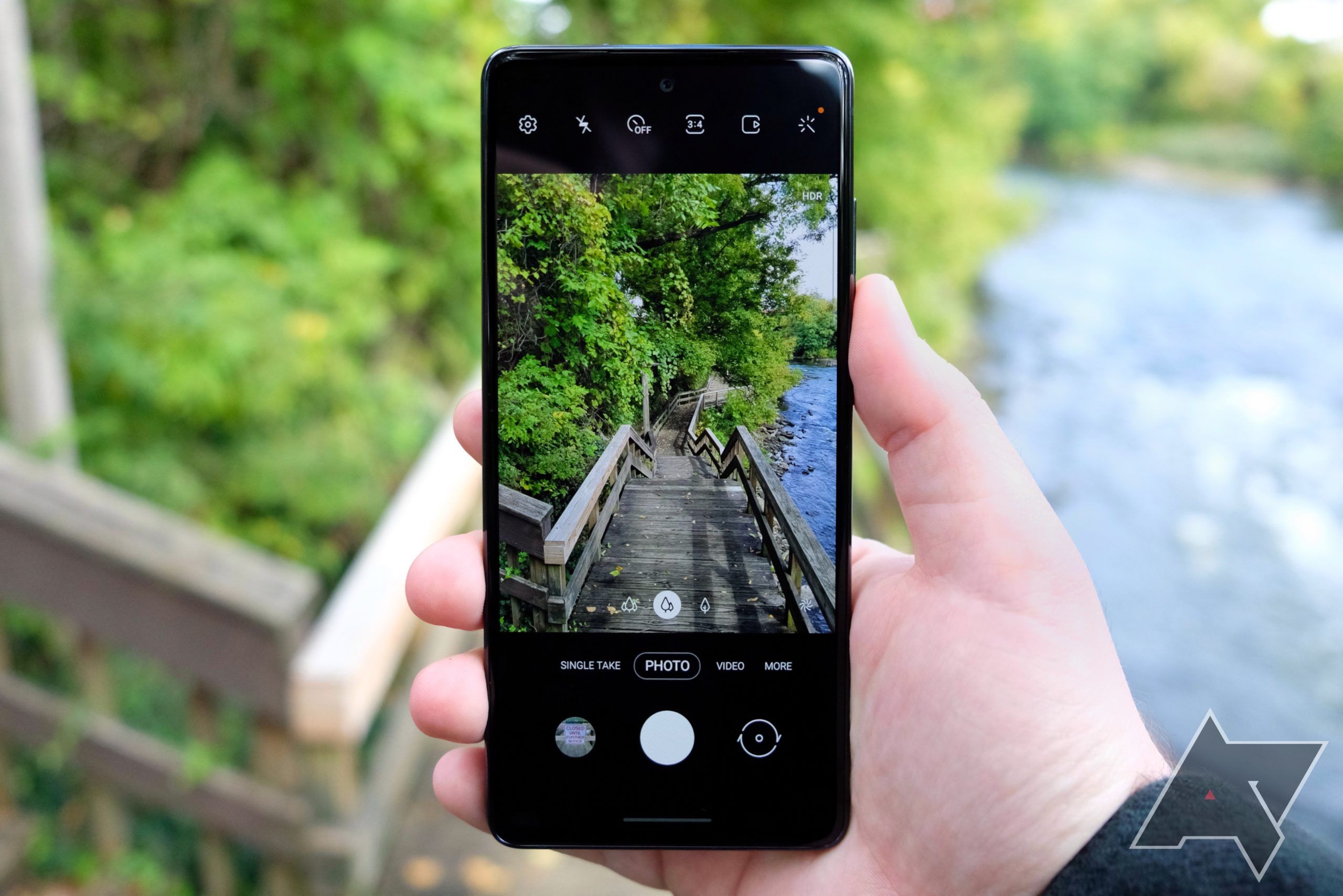 A person holding a Samsung Galaxy S20 FE with the camera app open with nature details in the background. 