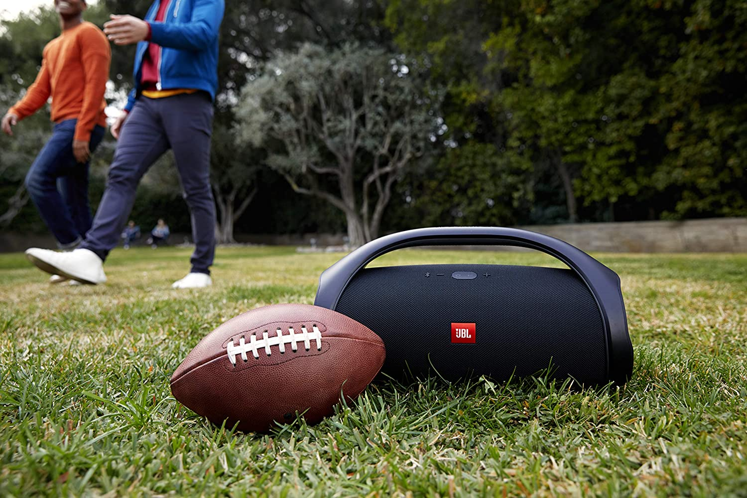 A JBL Boombox 2 in the grass with a football in front of it and people walking in the distance behind it.