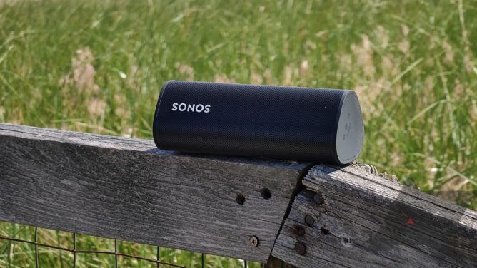 A Sonos Roam speaker placed on a wooden fence with grass in the background