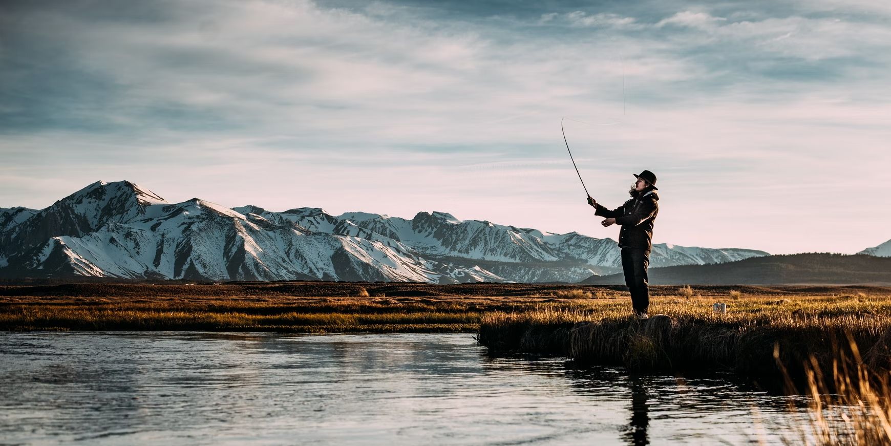 Um homem usando um chapéu de pesca verde-escuro está em pé na grama curta amarela, lançando uma vara de pescar em um lago cristalino cercado por paisagens montanhosas de tirar o fôlego.