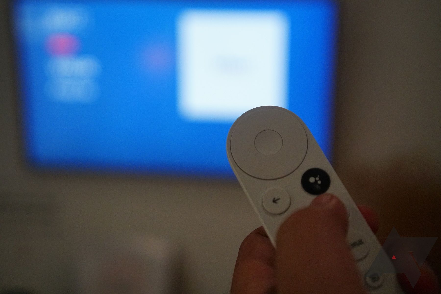 Hand using a white Google Chromecast remote with a blurry TV in the background.