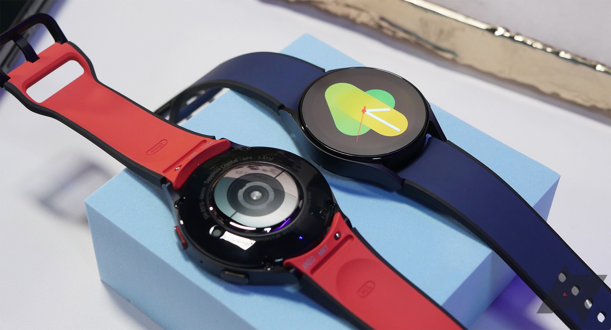 dua jam tangan pintar berada di blok biru pastel.  Satu memiliki tampilan yang menyala;  yang lainnya menghadap ke bawah. 