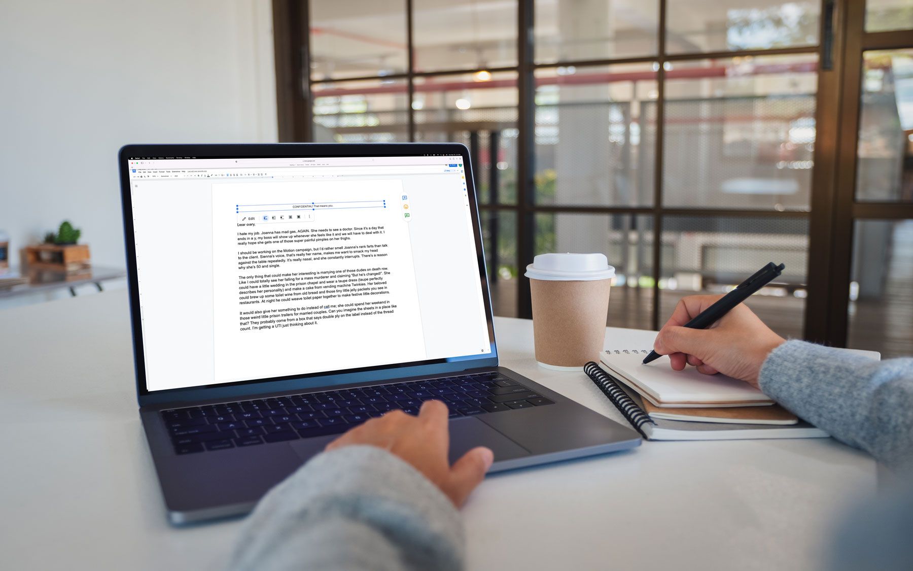 A person is sitting at a table with a cup of coffee and their laptop open. They're writing notes on a spiral pad with their right hand. 