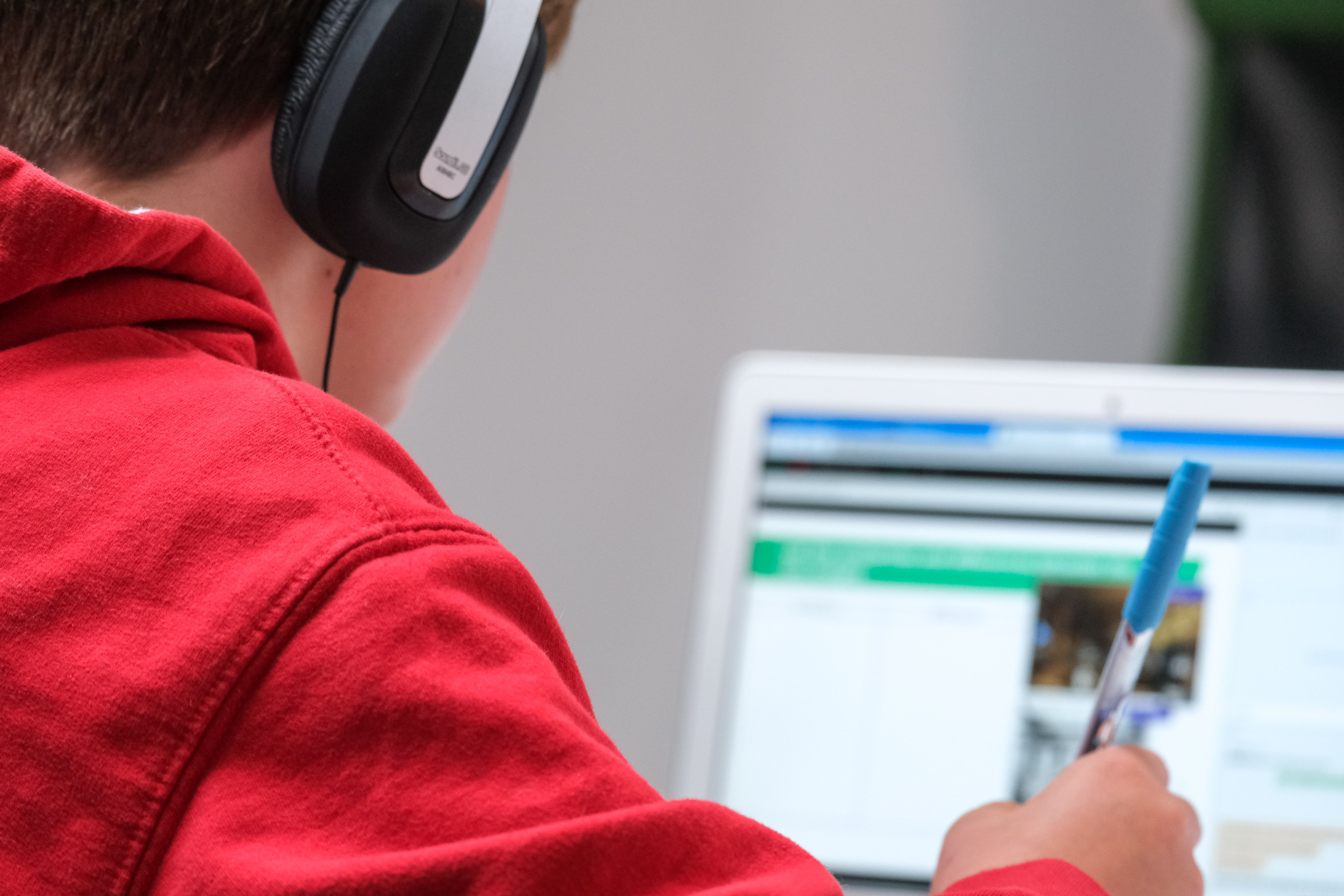 Child from the back wearing a red hoodie and headphones. The child has a pen in their right hand and a laptop is in the distance. 