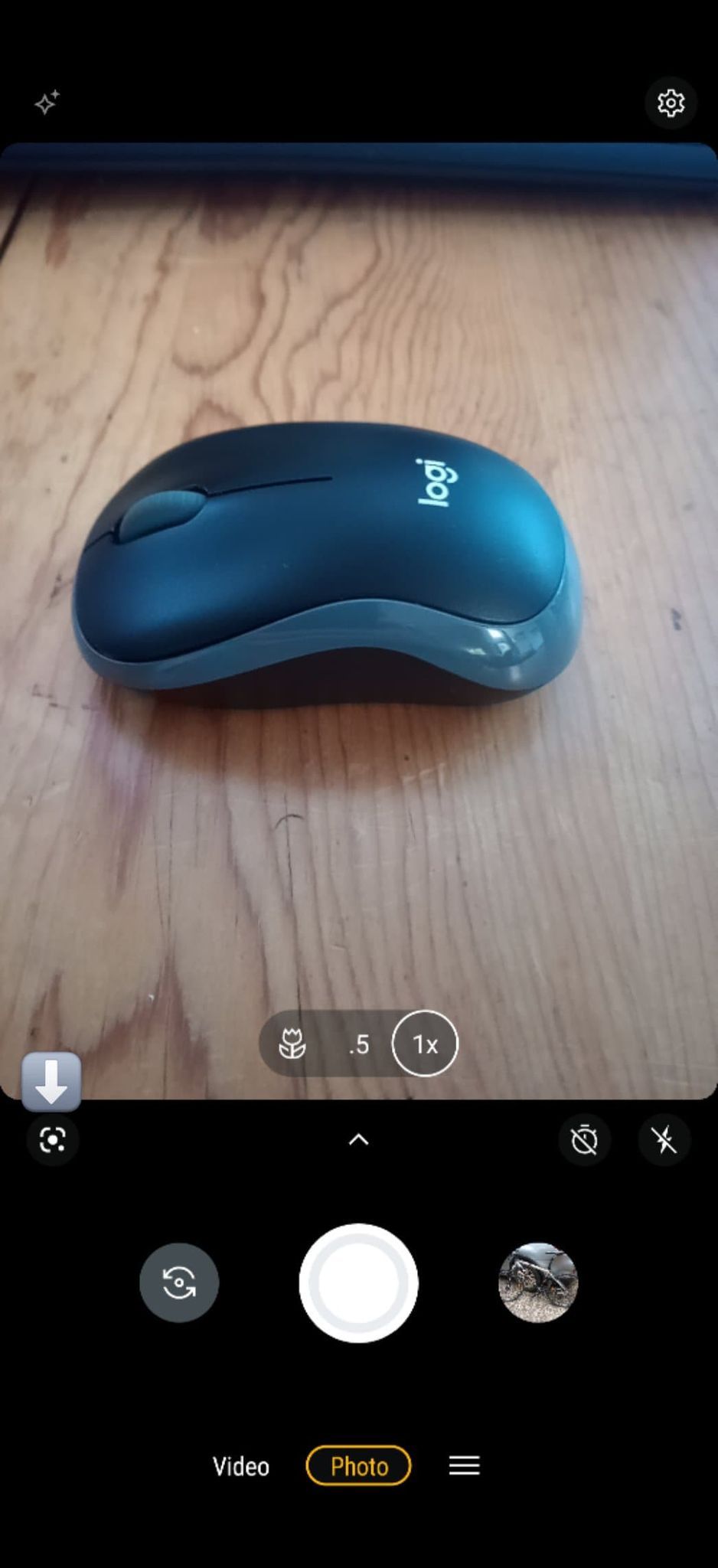 Image shows the Android camera app open with a computer mouse in the display and an arrow pointing to the Google Lens icon in the lower left.