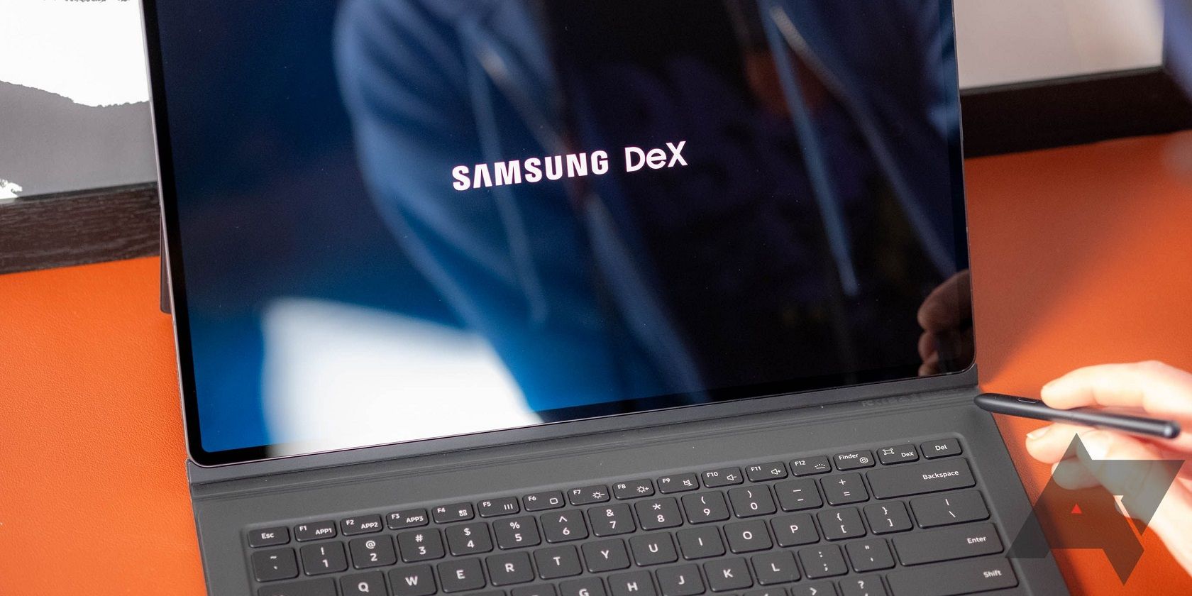 Image shows a tablet with a keyboard open displaying the words Samsung DeX, and a person's hand holding a stylus.