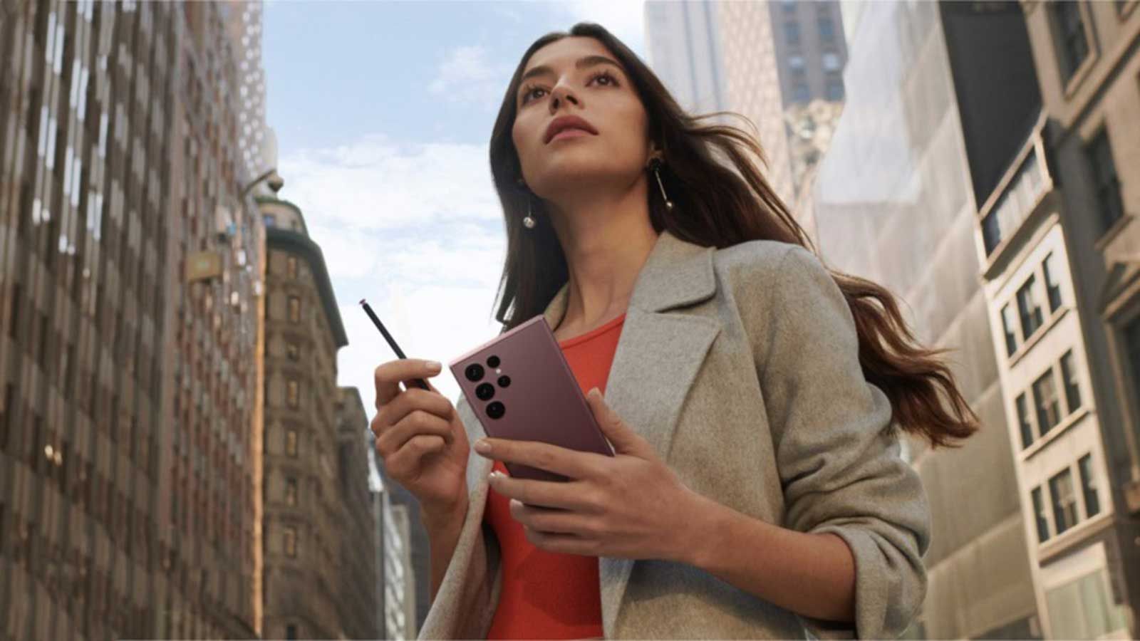 Wanita yang berdiri di kota besar melihat ke depan sambil memegang telepon ungu dan stylus. 