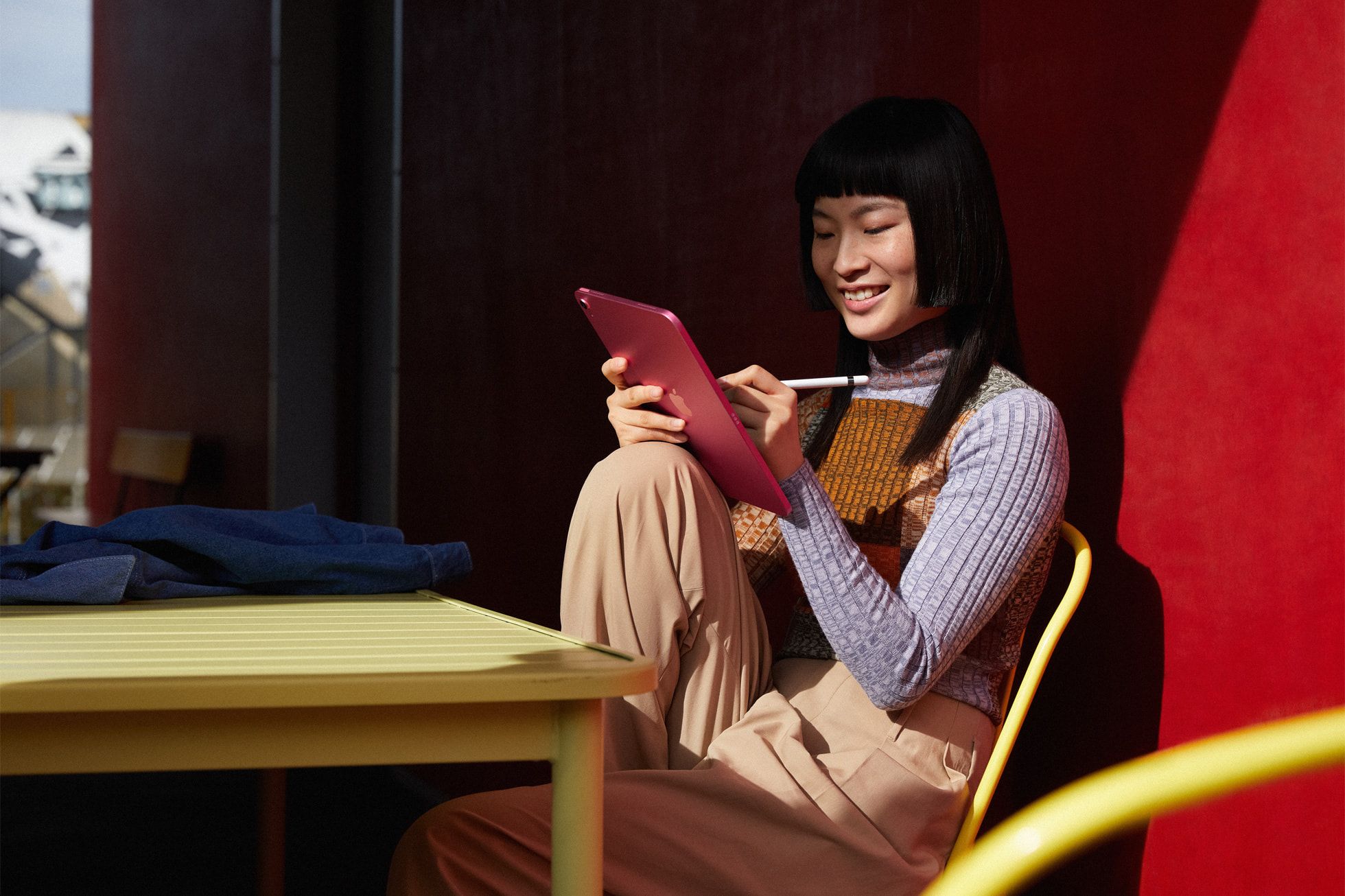 A person sits in a chair with colorful red background holding an iPad while drawing into it and smiling.