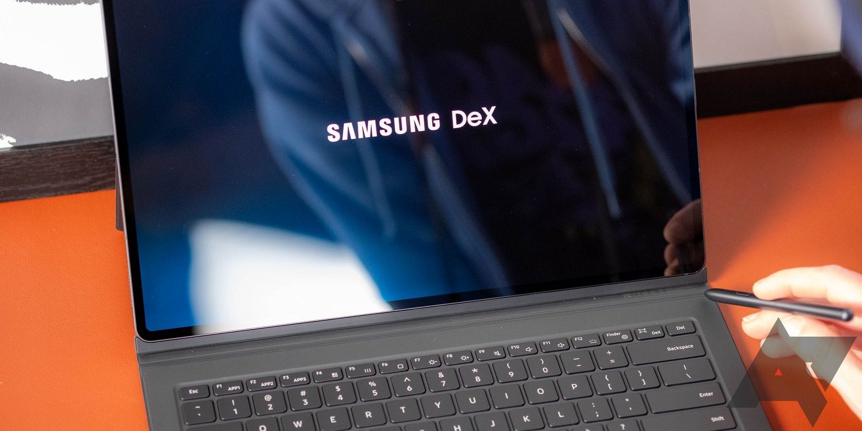 Image shows a Samsung tablet with a keyboard attached, open, with the words Samsung DeX displayed on the screen. A hand is holding a stylus in the lower right corner toward the screen.