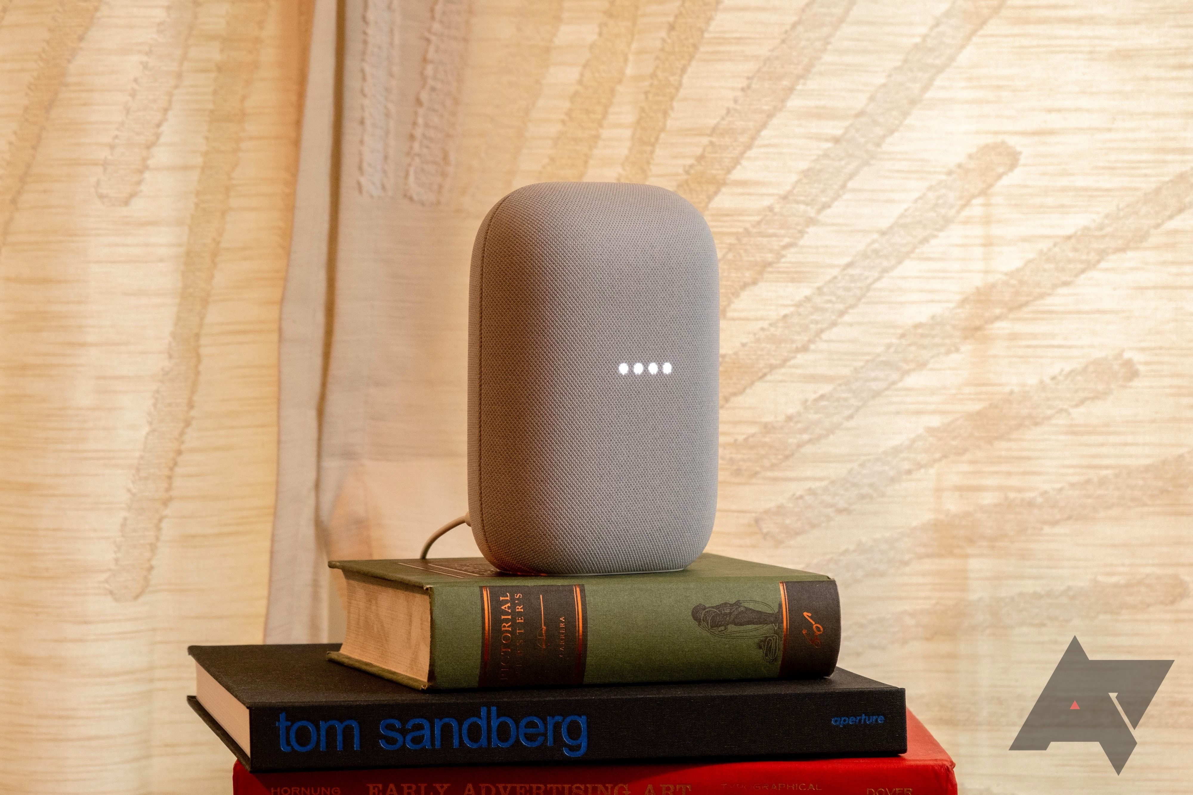 A Grey Google Nest Audio on a stack of books with a yellow curtain background.