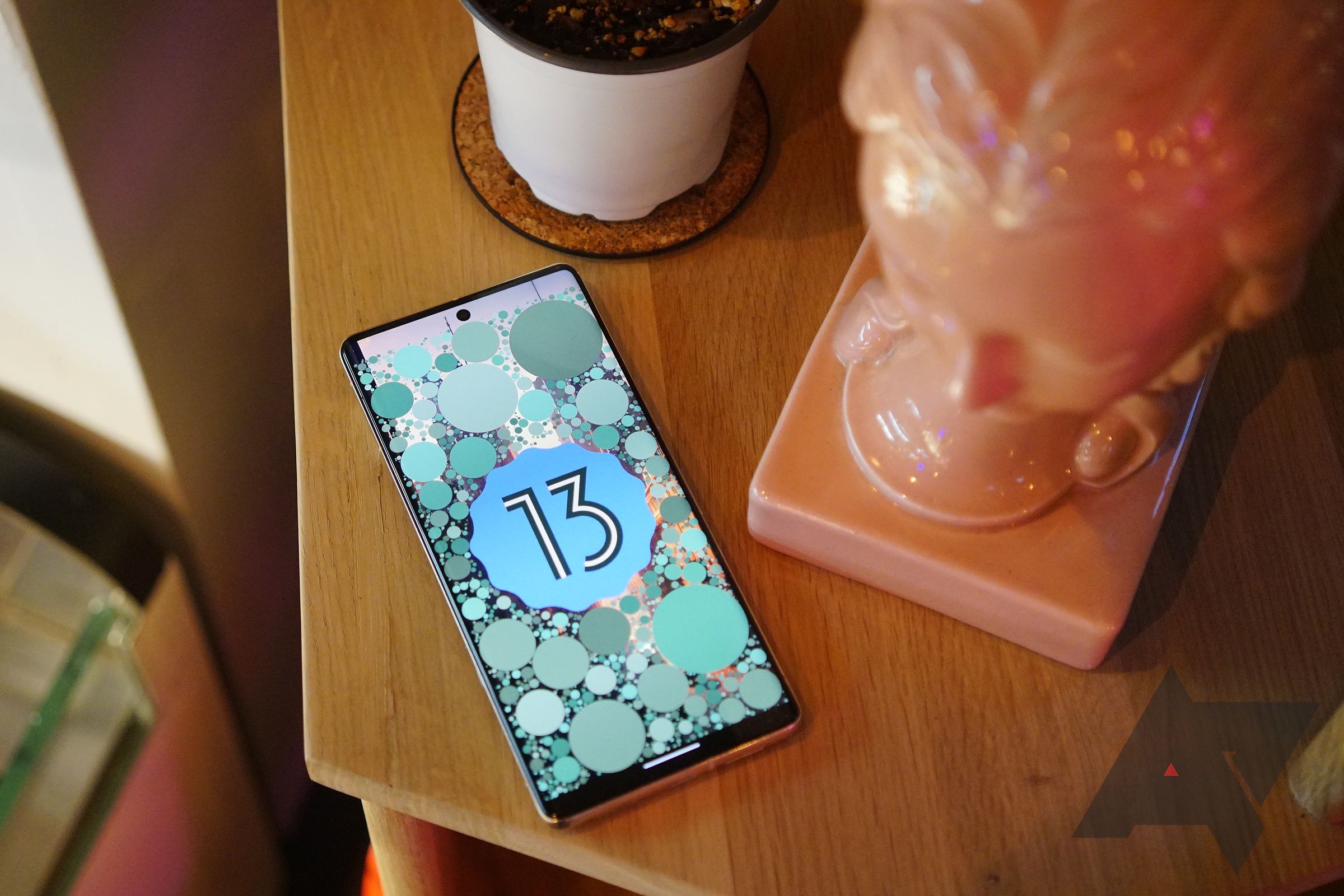 A phone with an illuminated display with the number "13" sits on a teak table. A pink bust and seedling are placed next to it. 