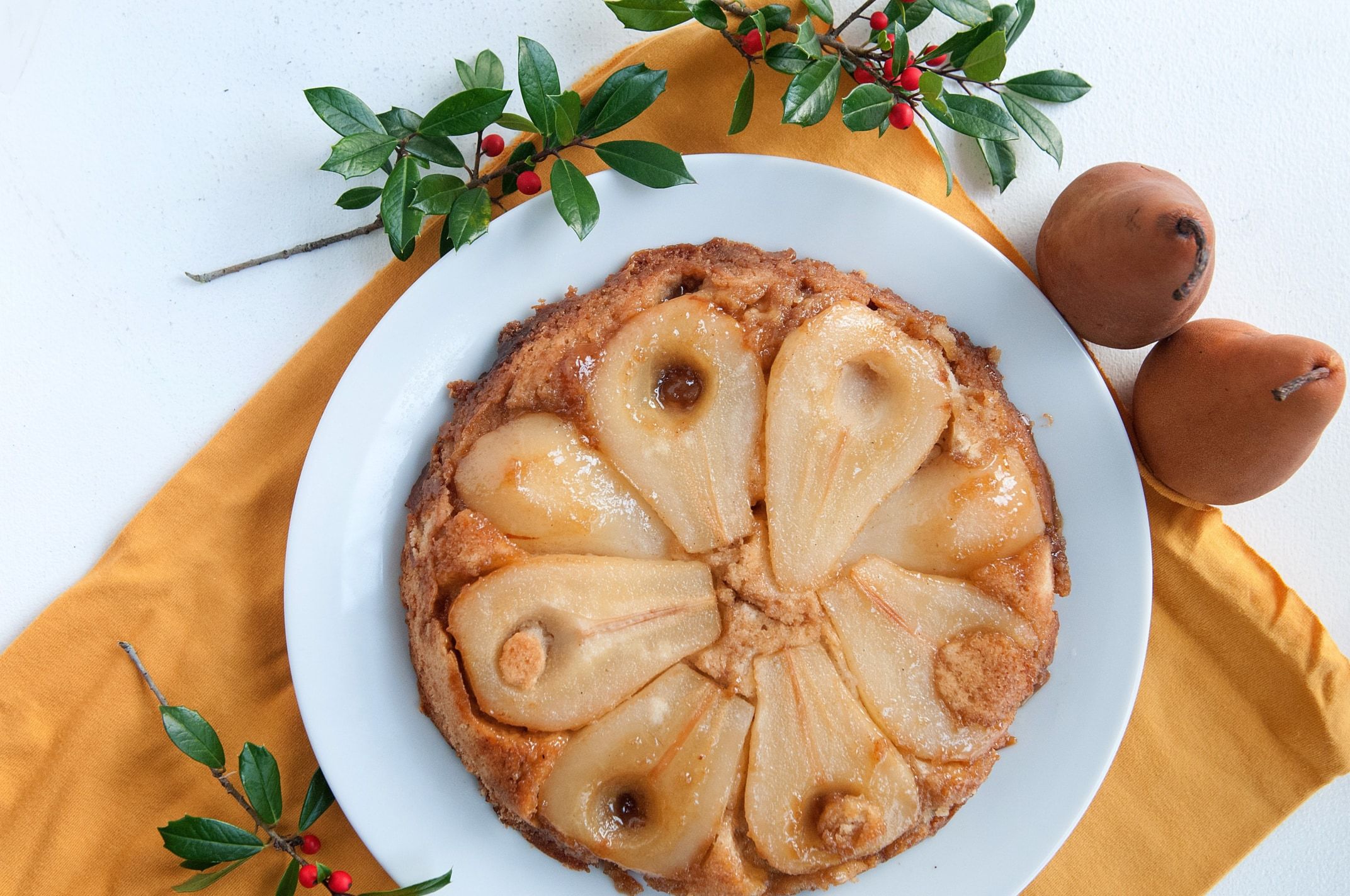 a pear upside down cake sits on a wooden platter and is surrounded by holly and pears. 