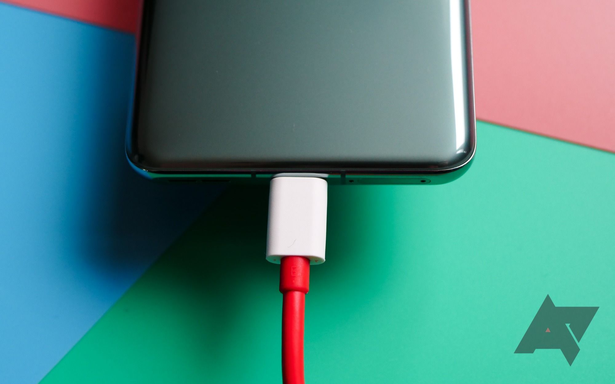 The rear bottom third of a OnePlus 11 smartphone hovers over a tri-colored cardboard background with a white and red USB-C cable plugged in