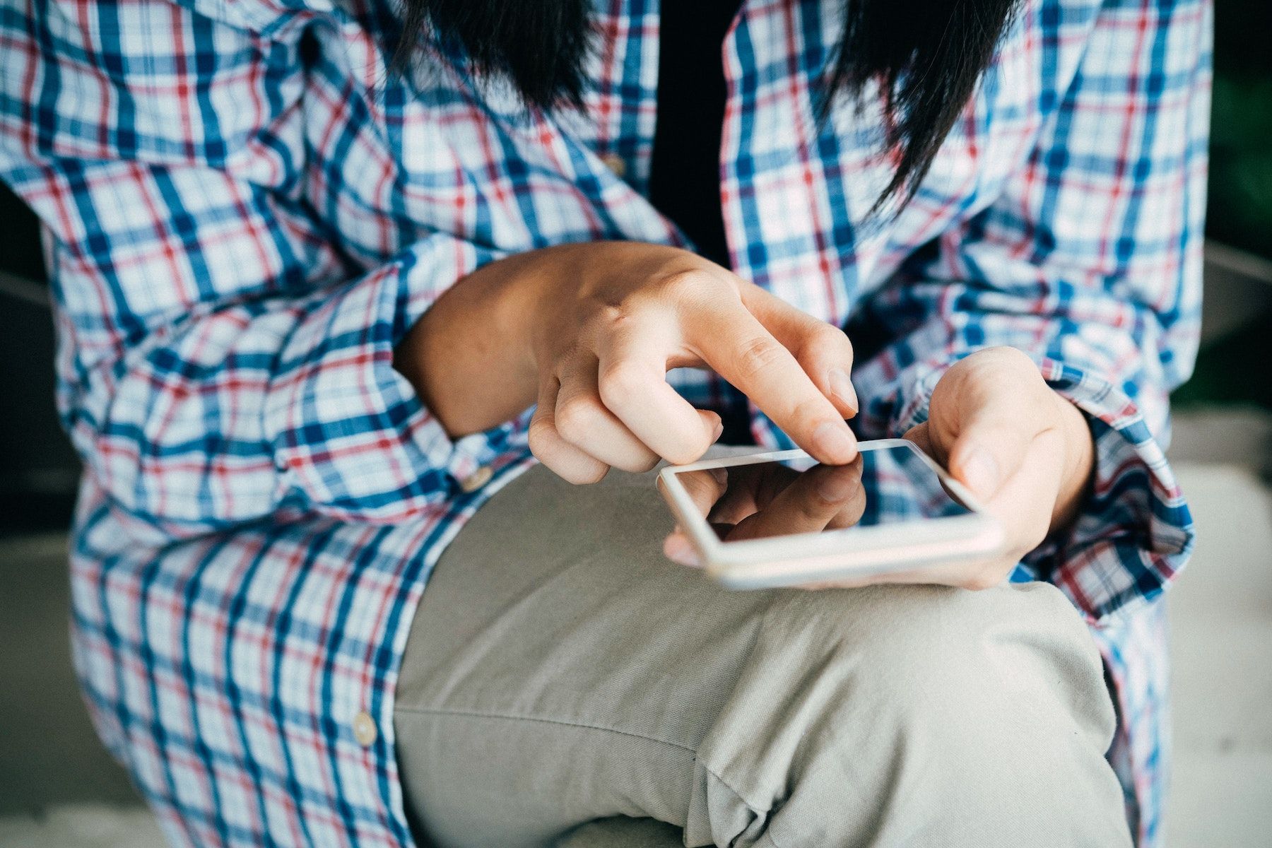 A person in a plaid shirt and khakis holds a phone in their left hand while their right index finger taps the screen. 