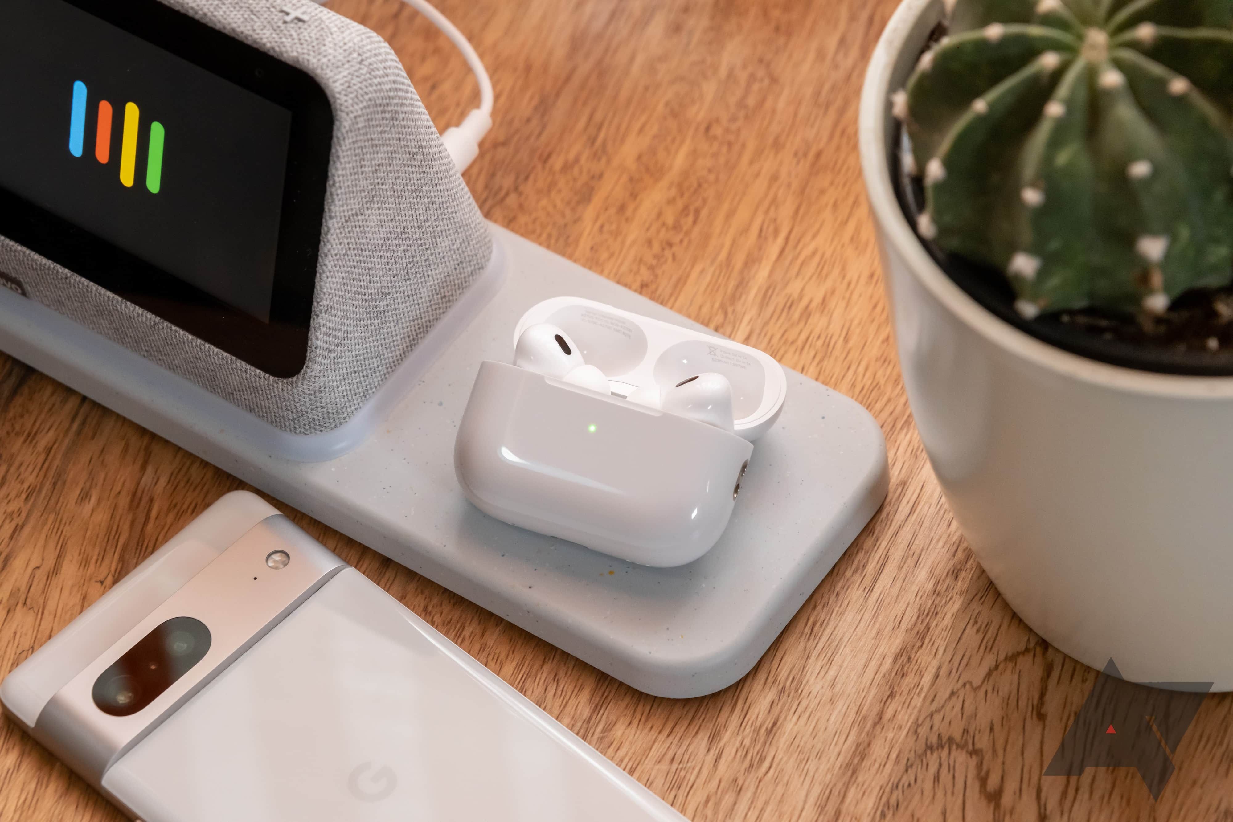 Wireless earbuds in their charging case on a table with a smartphone, a smart display, and a small cactus.