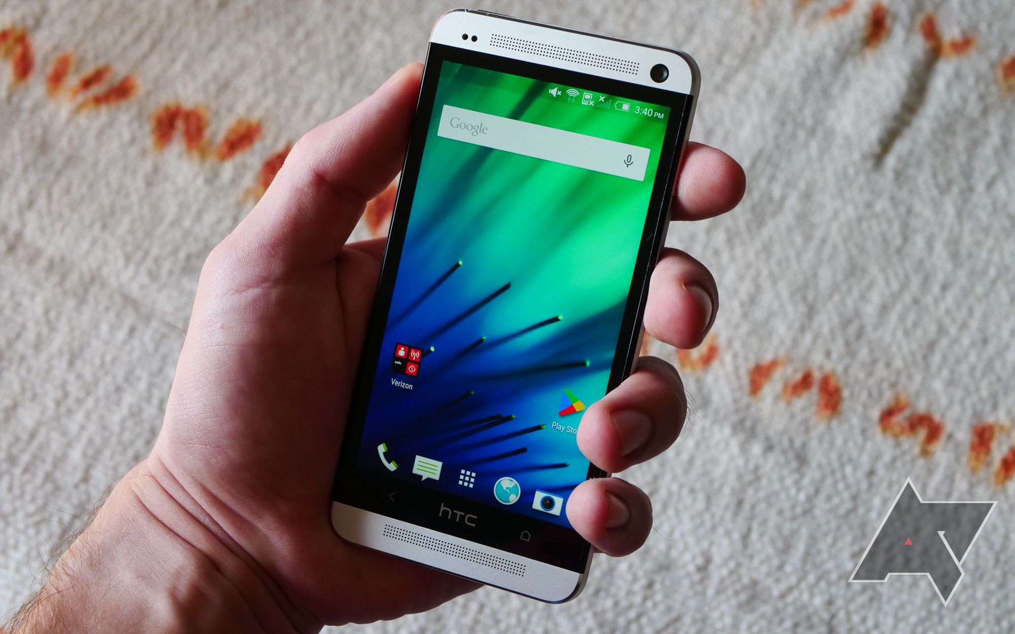 A man's hand holds an HTC M7 with a blue and green screen.
