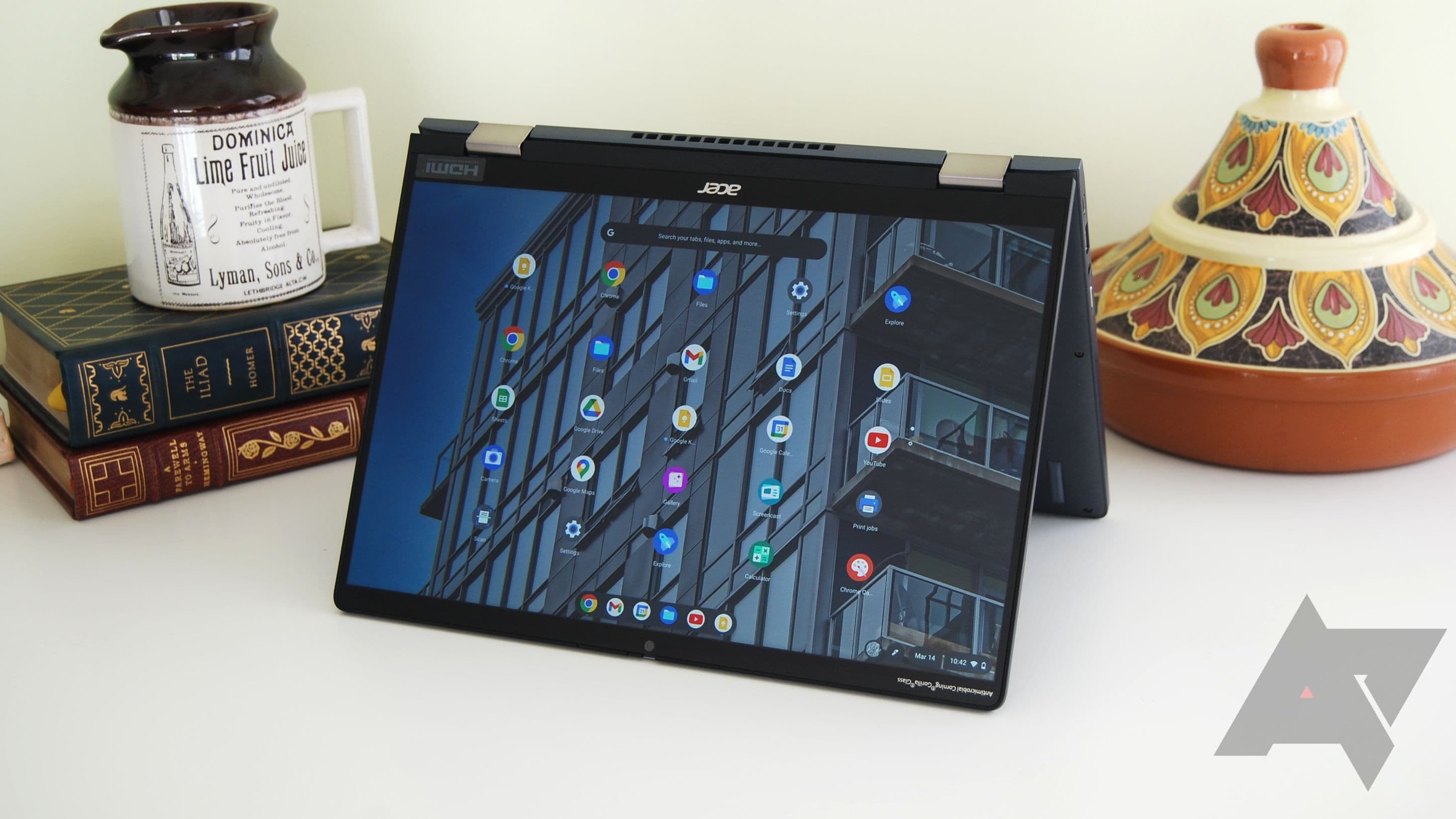 An Acer Chromebook Spin 714 in tent mode on a white table with books, a mug, and a piece of pottery in the background