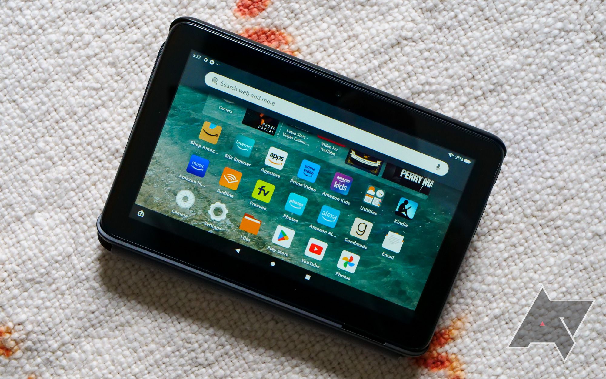 An Amazon Fire 7 tablet sitting on a blanket with the home screen displayed.