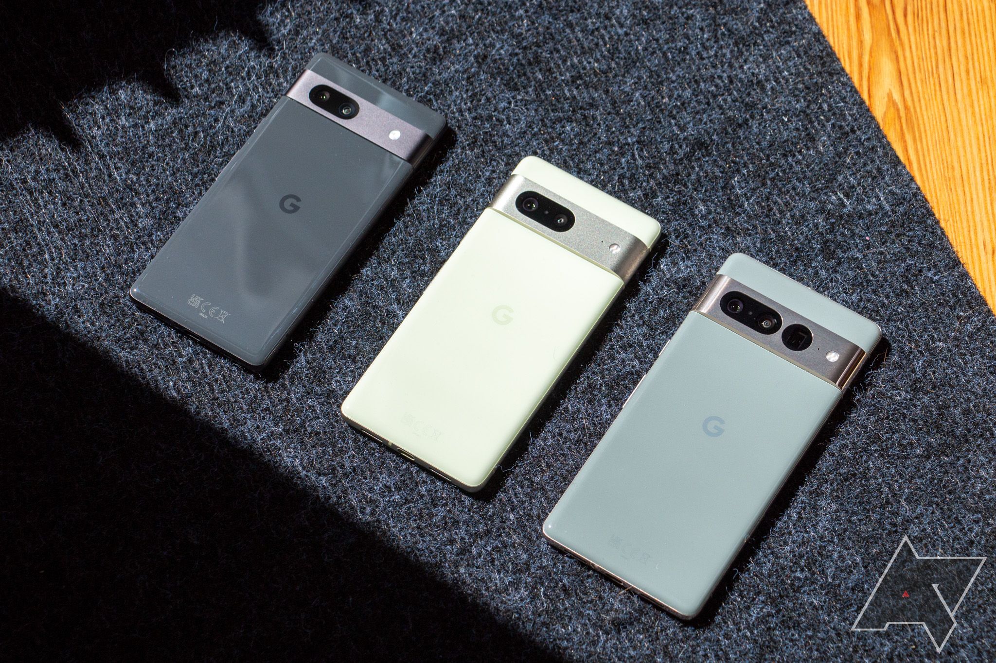 Google Pixel 7a, Pixel 7, and Pixel 7 Pro lying face-down next to each other on dark gray textured surface in sunlight.