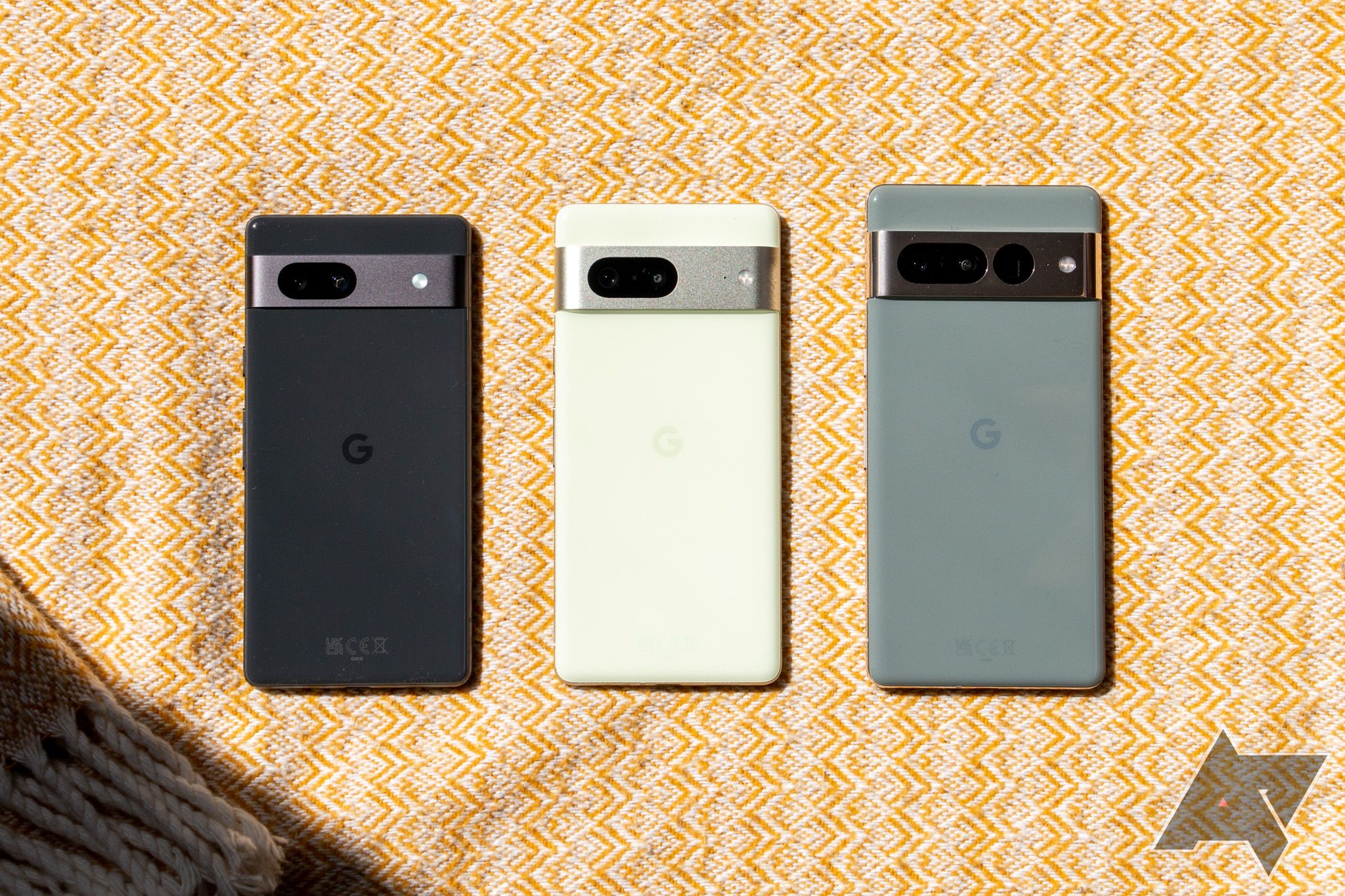 The Google Pixel 7a, Pixel 7, and Pixel 7 Pro lying neatly in a line face down on a patterned yellow surface