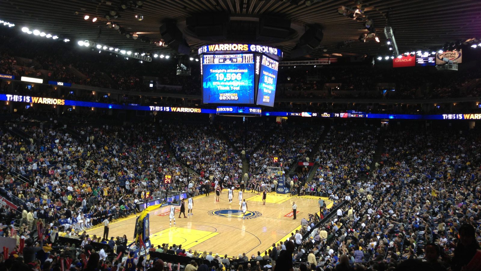 A packed arena watching a basketball game. The large display above the court names the Warriors as one of the teams playing.