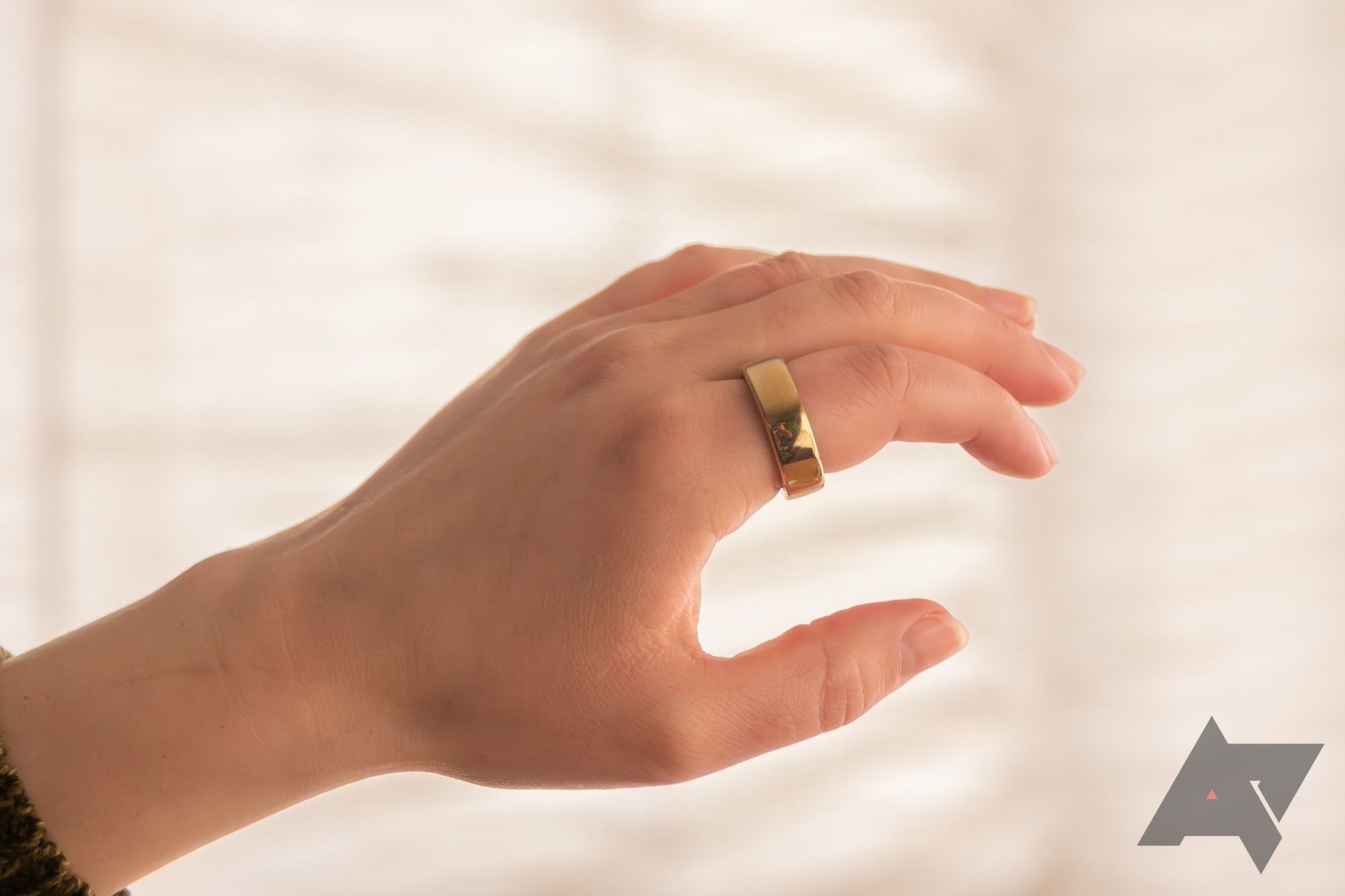 A golden Oura Ring Gen 3 being worn on the index finger over an off-white background
