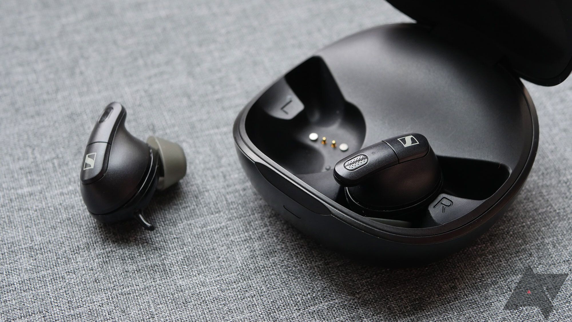 Sennheiser Conversation Clear Plus earbuds, with one of them inside their charging case and the other lying next to it