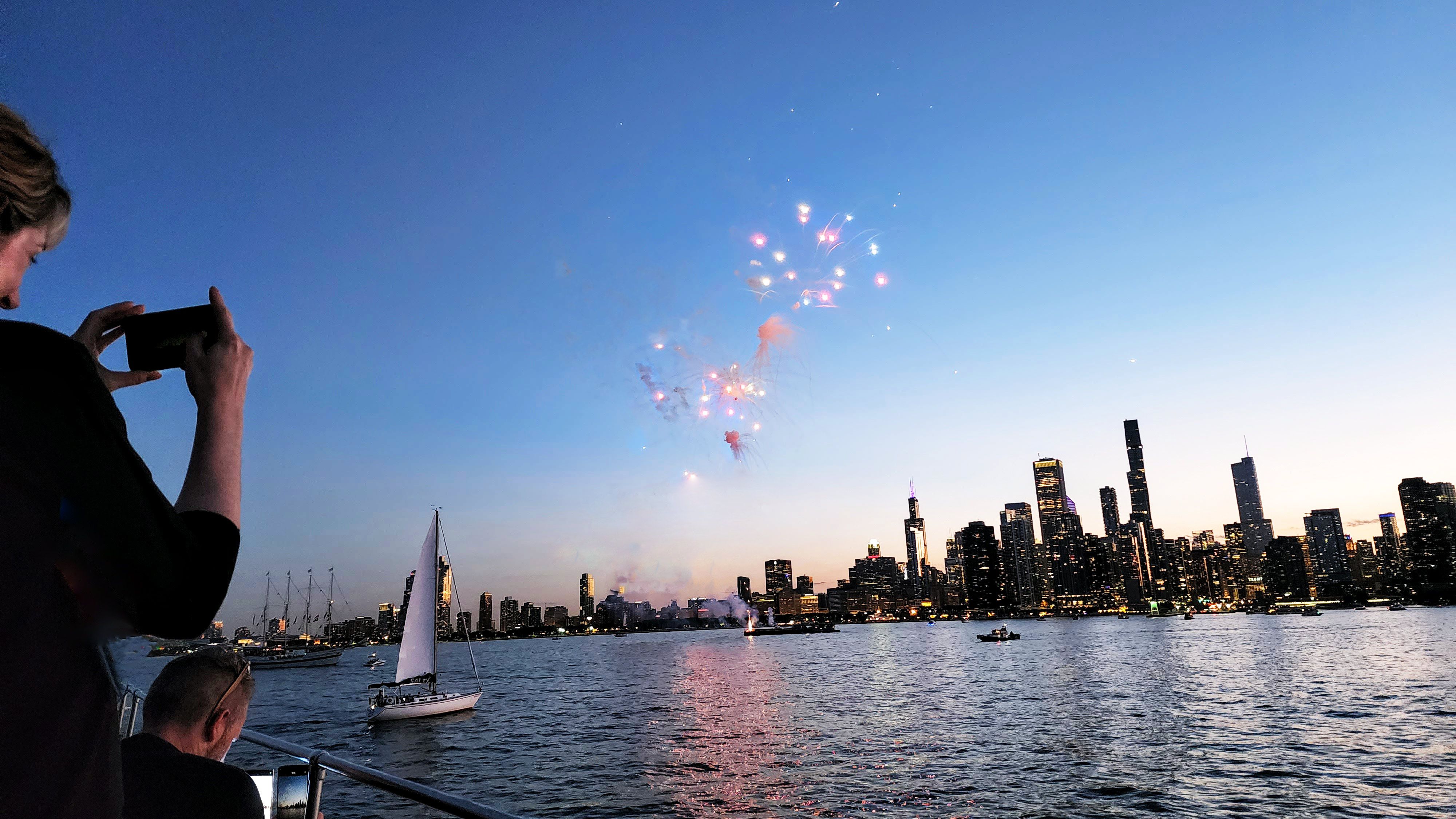 Two people on a boat using their smartphones to take photos of fireworks over the Chicago skyline. 