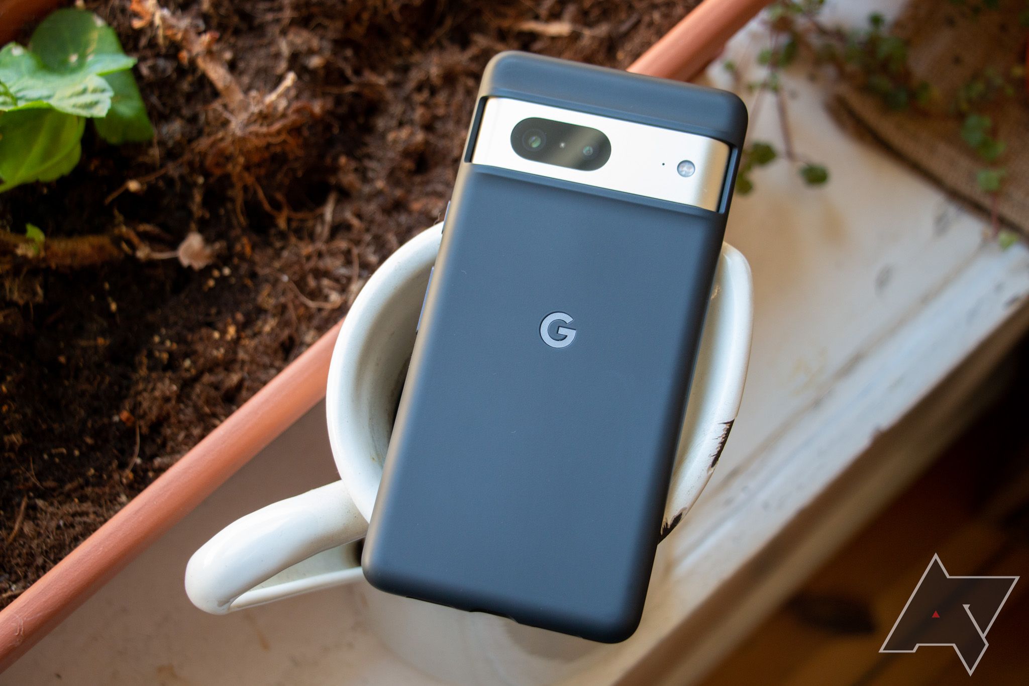 A Google Pixel 7 in a black first-party Google case lying face-down on the top of a watering can amidst flower pots