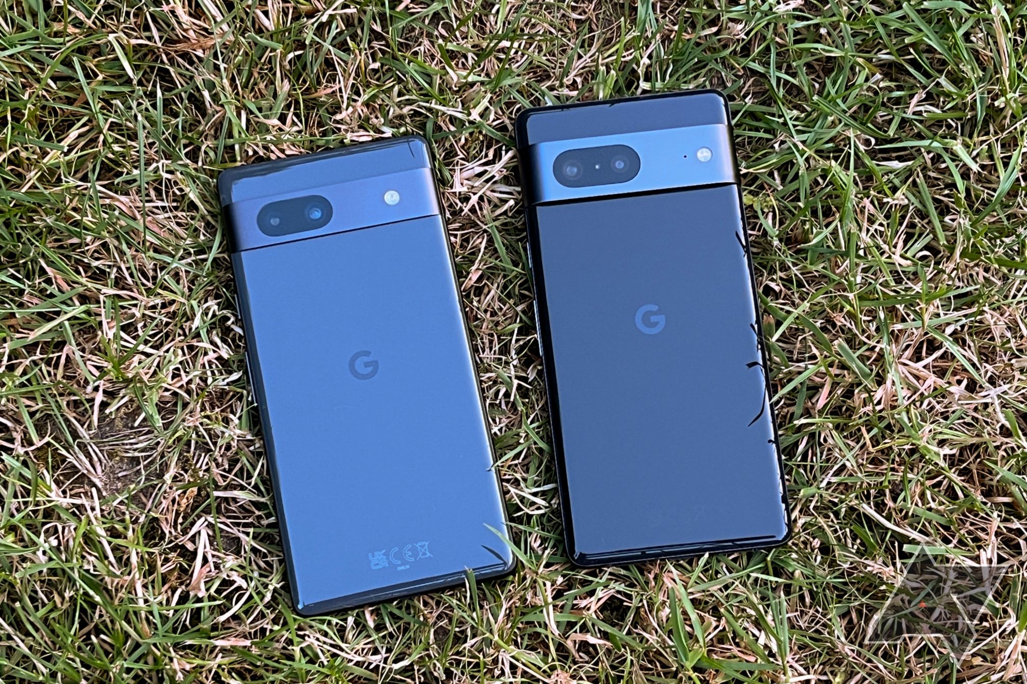 A black Google Pixel 7a and a black Pixel 7 lying display-down on grass next to each other