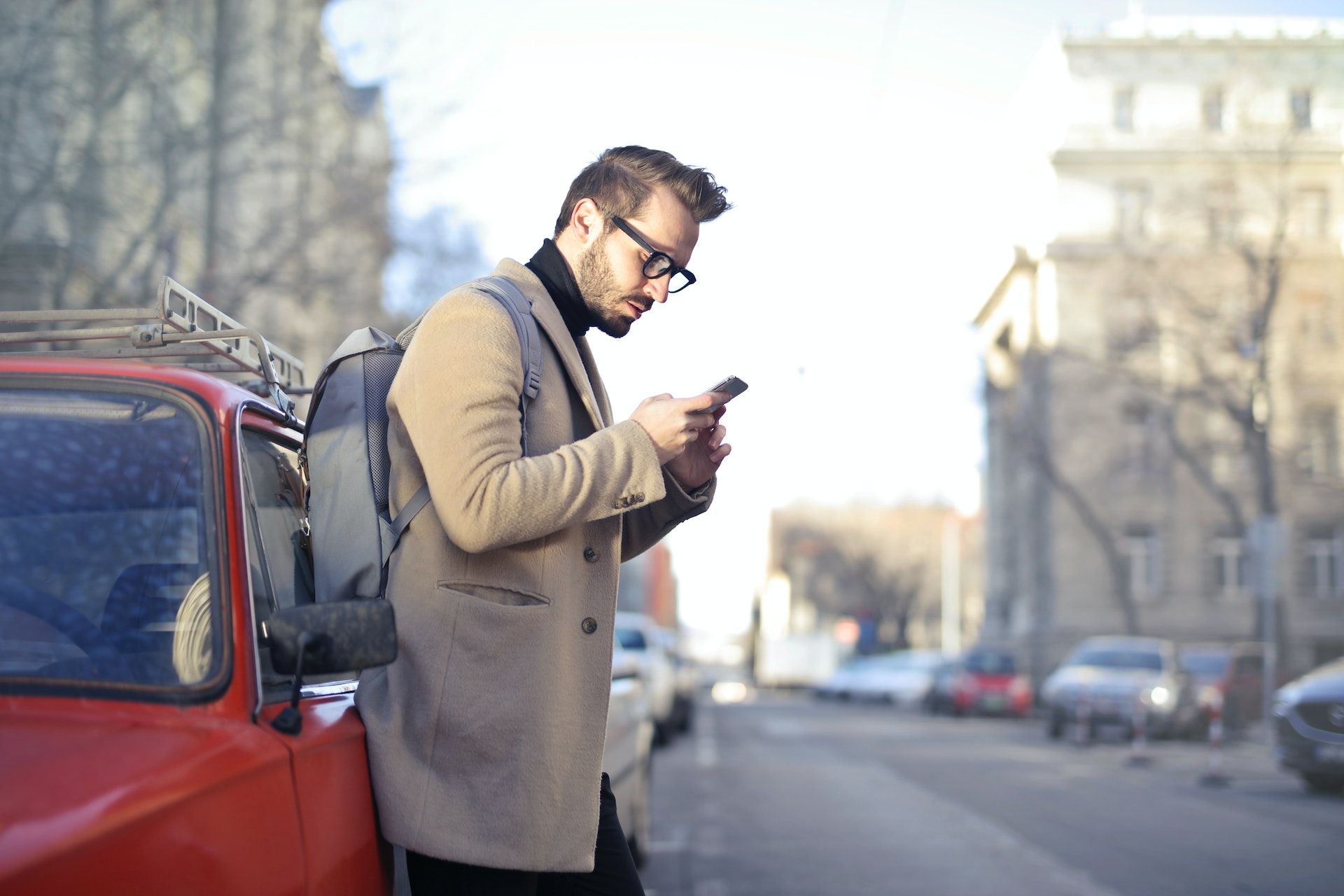 Man in Beige Coat Holding Phone Leaning on Red Vehicle