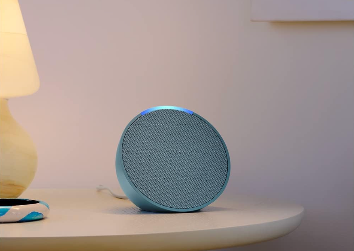 A blue Amazon Echo pop on a nightstand next to a lamp.