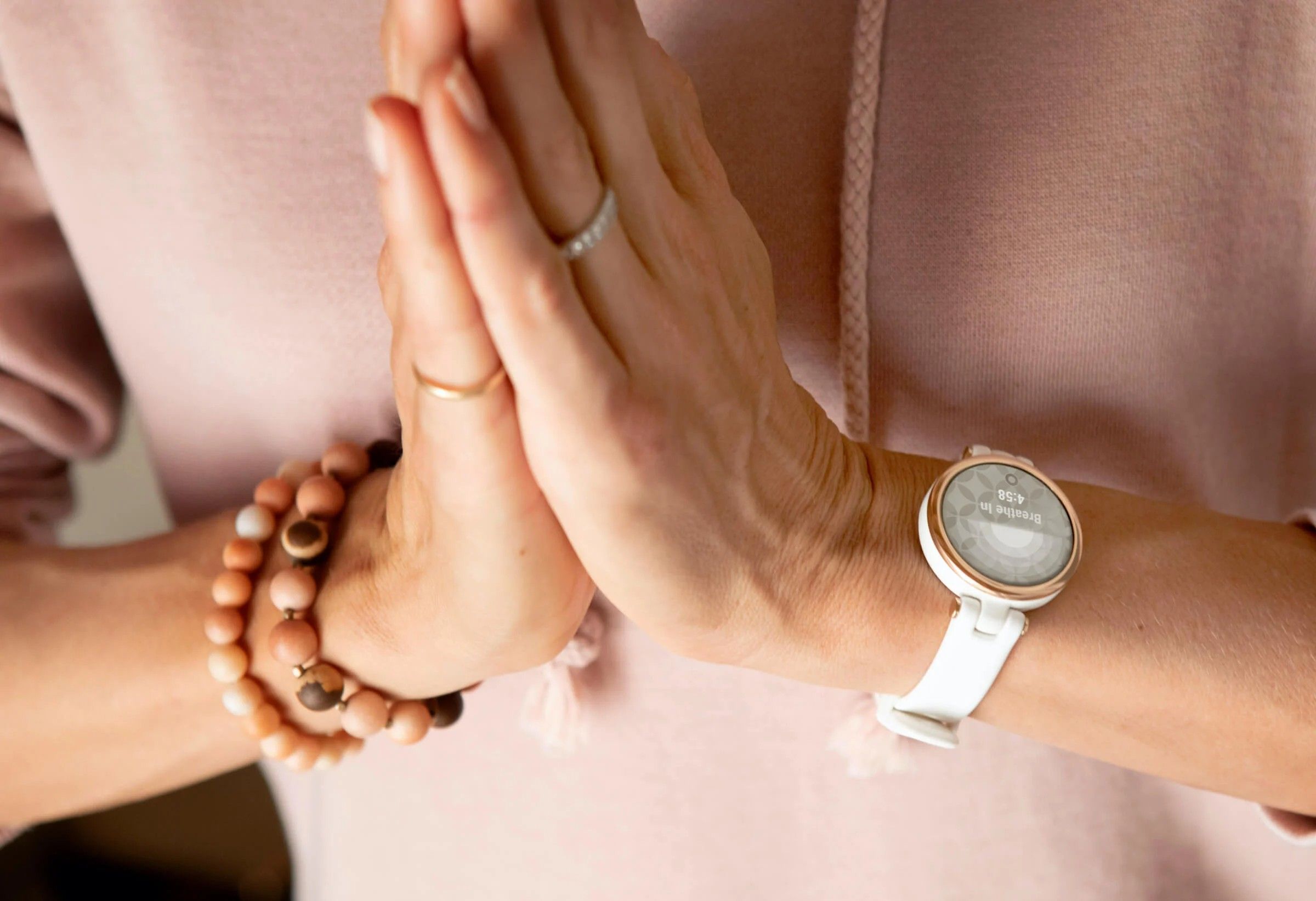 An individual with a pink sweater practicing a mindfulness exercise with hands in a prayer position, wearing a white smartwatch