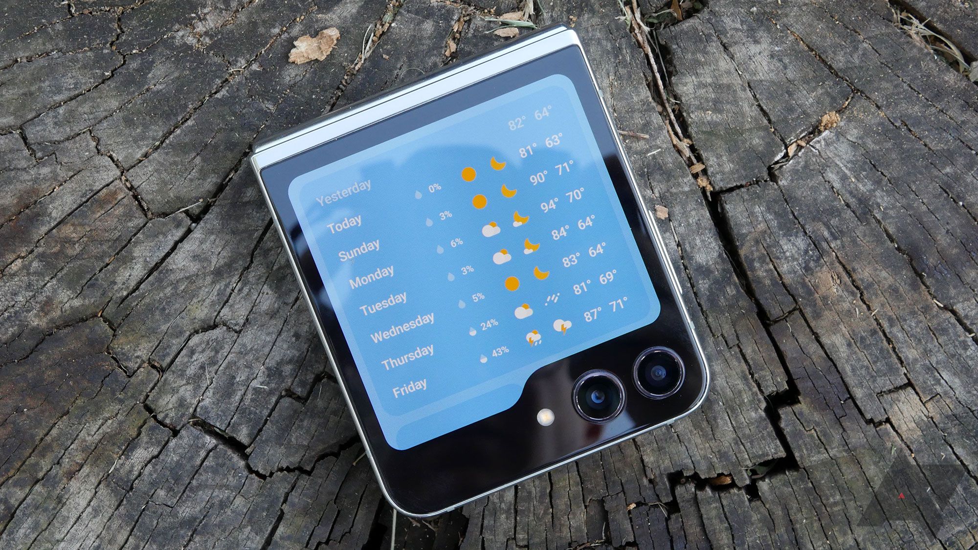 Samsung's Galaxy Z Flip 5 laying closed on tree trunk wood with the weather widget showing.
