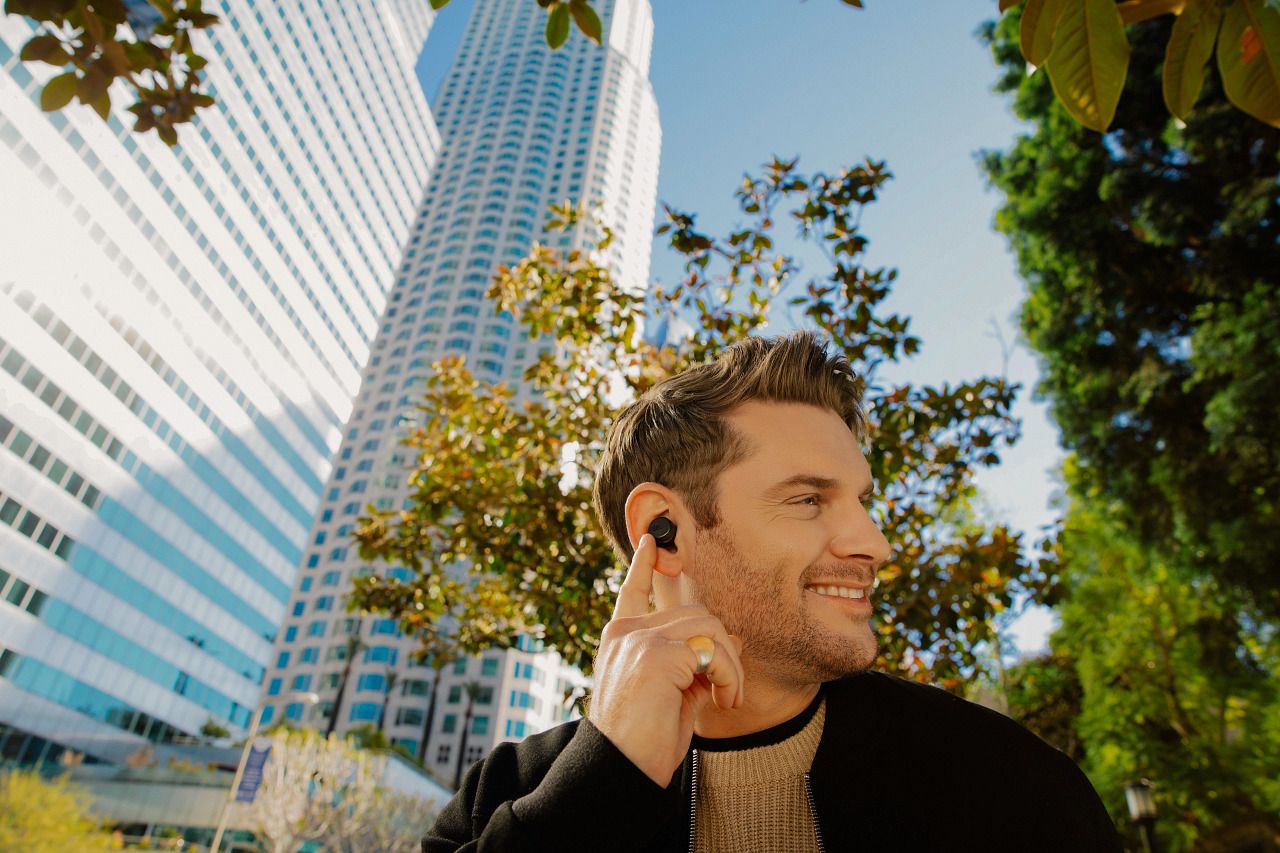 A person wearing the JBL Tour Pro+ earbuds and pointing at them against an outdoor background.