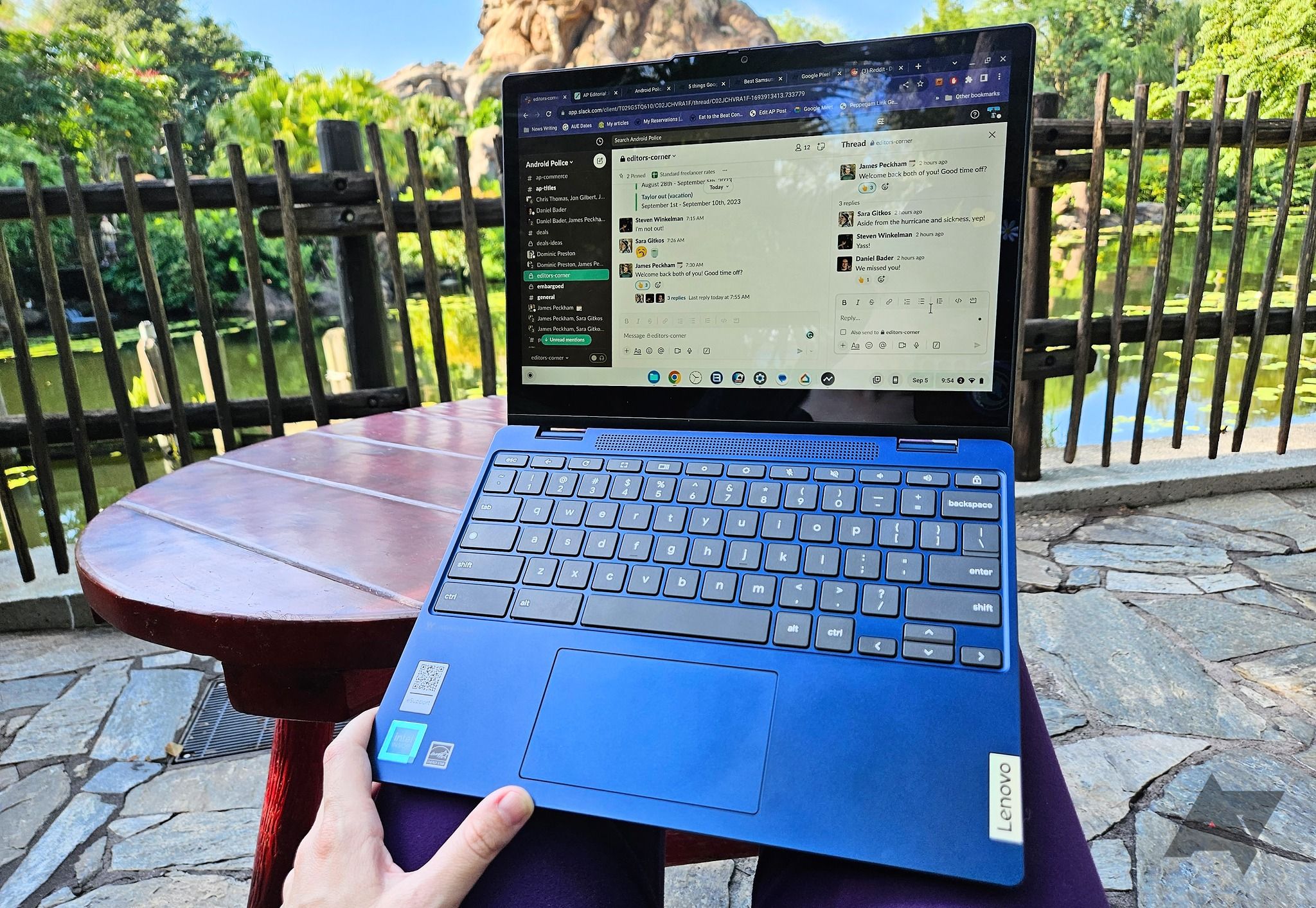 A Lenovo Flex 3i Chromebook (Gen 8) on someone's knees while sitting outside on a stone patio with a pond with greenery around it in front of them