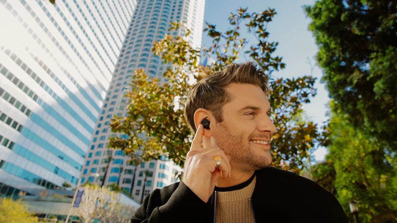 A person wearing the JBL Tour Pro+ earbuds and pointing at them against an outdoor background