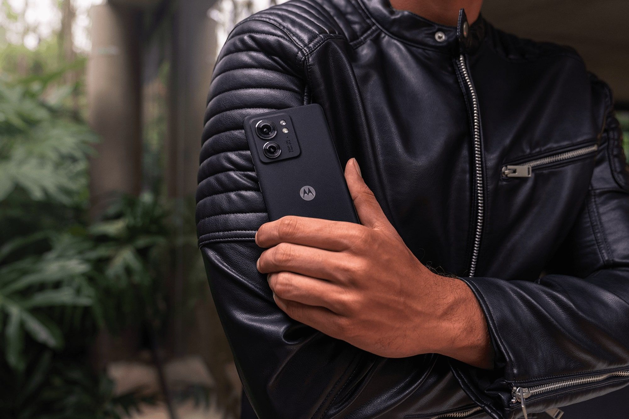 Motorola edge (2023) held by a person in a leather jacket against a forested background 