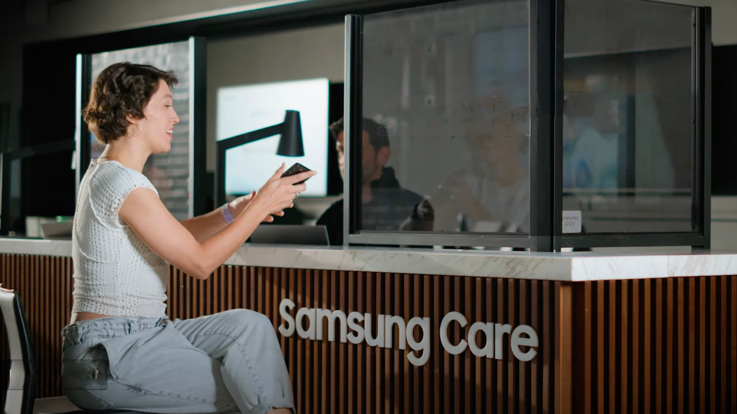 A woman sits at a Samsung Care counter holding a phone
