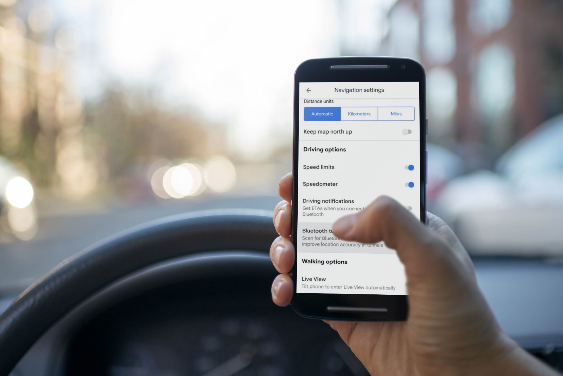 A hand holding a phone in a car with a screenshot of the Google Maps settings page shown on the phone
