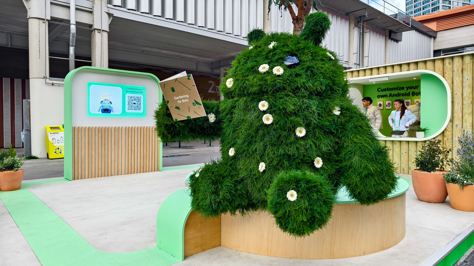 An Android plant robot statue covered in grass and flowers holding a 'Gardening for Bots' book at Google's Android Avenue in MWC 2024