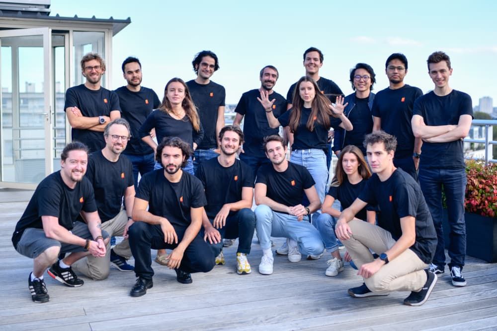 A team photo of the Mistral AI group on a rooftop. They are wearing matching black t-shirts with an orange logo, displaying a mix of standing and seated poses, with a city skyline in the background.
