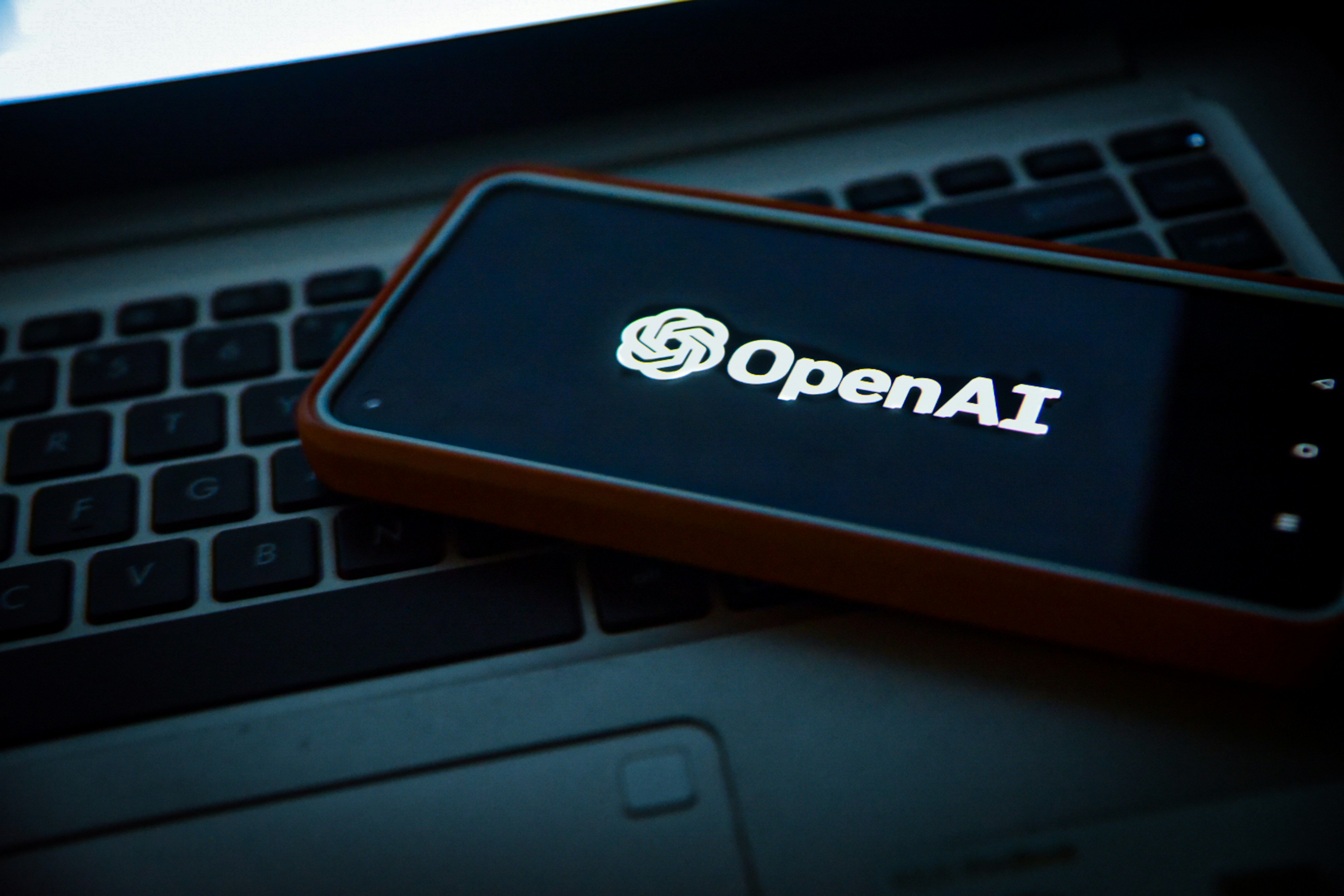 A smartphone with the OpenAI logo on the screen rests on a laptop keyboard.