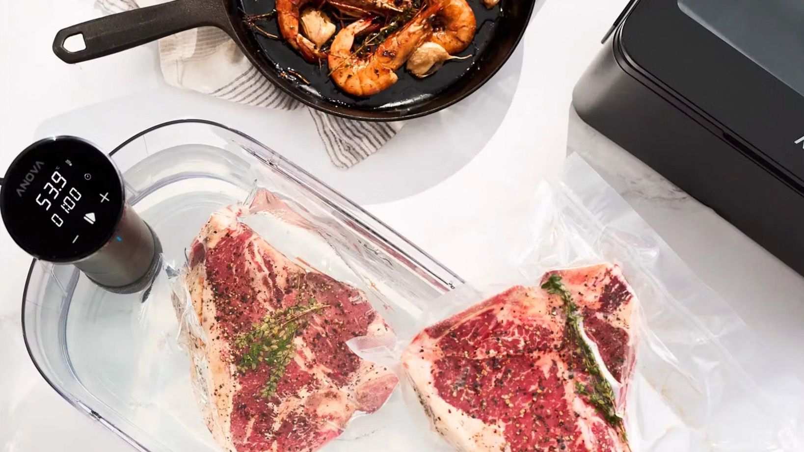 Photo of an Anova smart sous vide cooking two sealed steaks, with prawns in a pan nearby