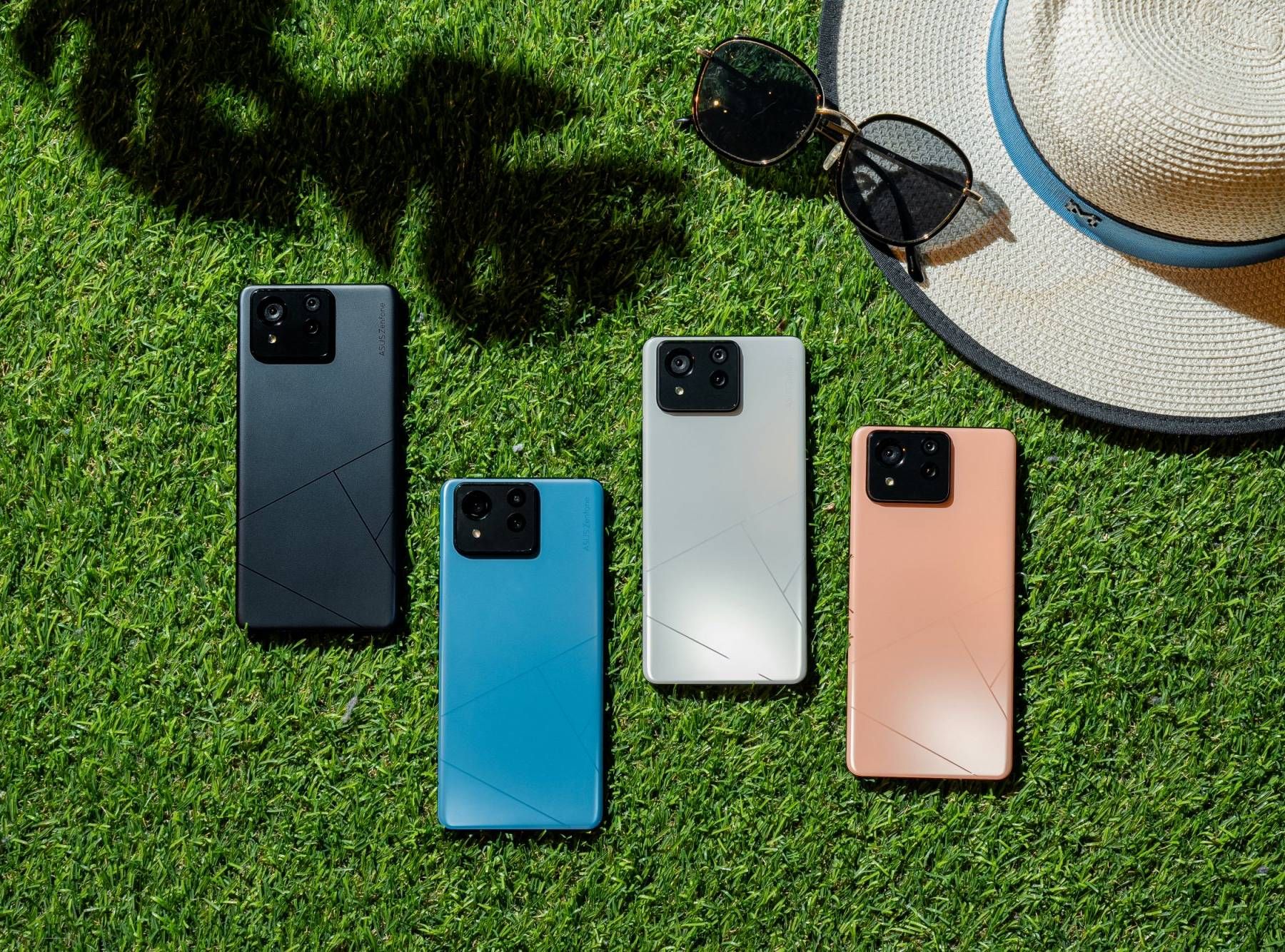 Four different colors of the Asus Zenfone 11 Ultra sitting face down on the grass nearby a sun hat and sunglasses