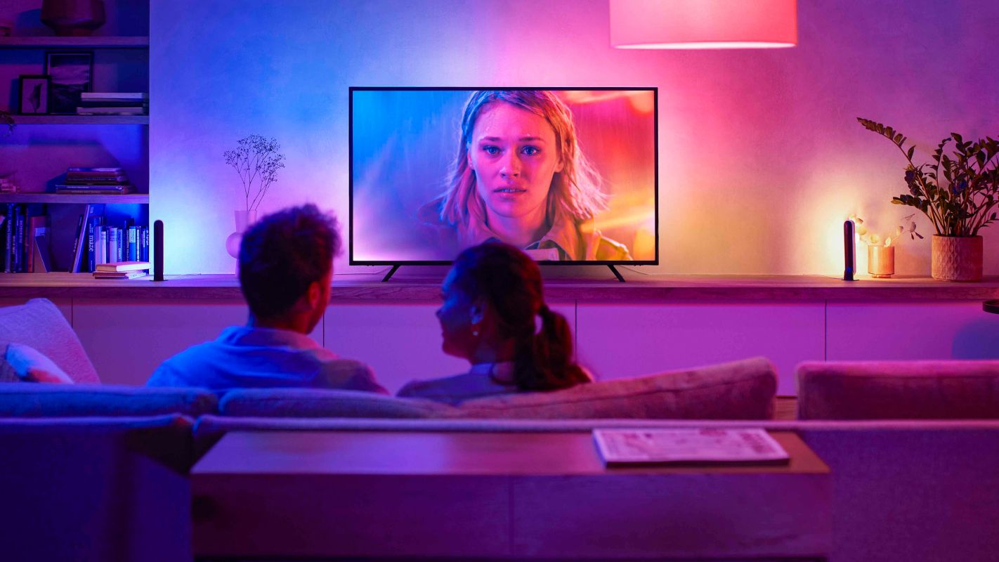 A couple sitting on a couch with their backs to the camera, watching TV with ambient lighting matching the content on the screen