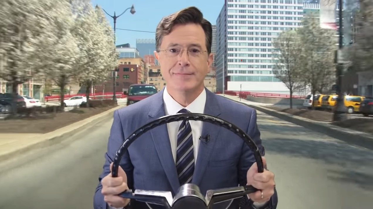 A screen capture of Stephen Colbert driving an imaginary car through the city