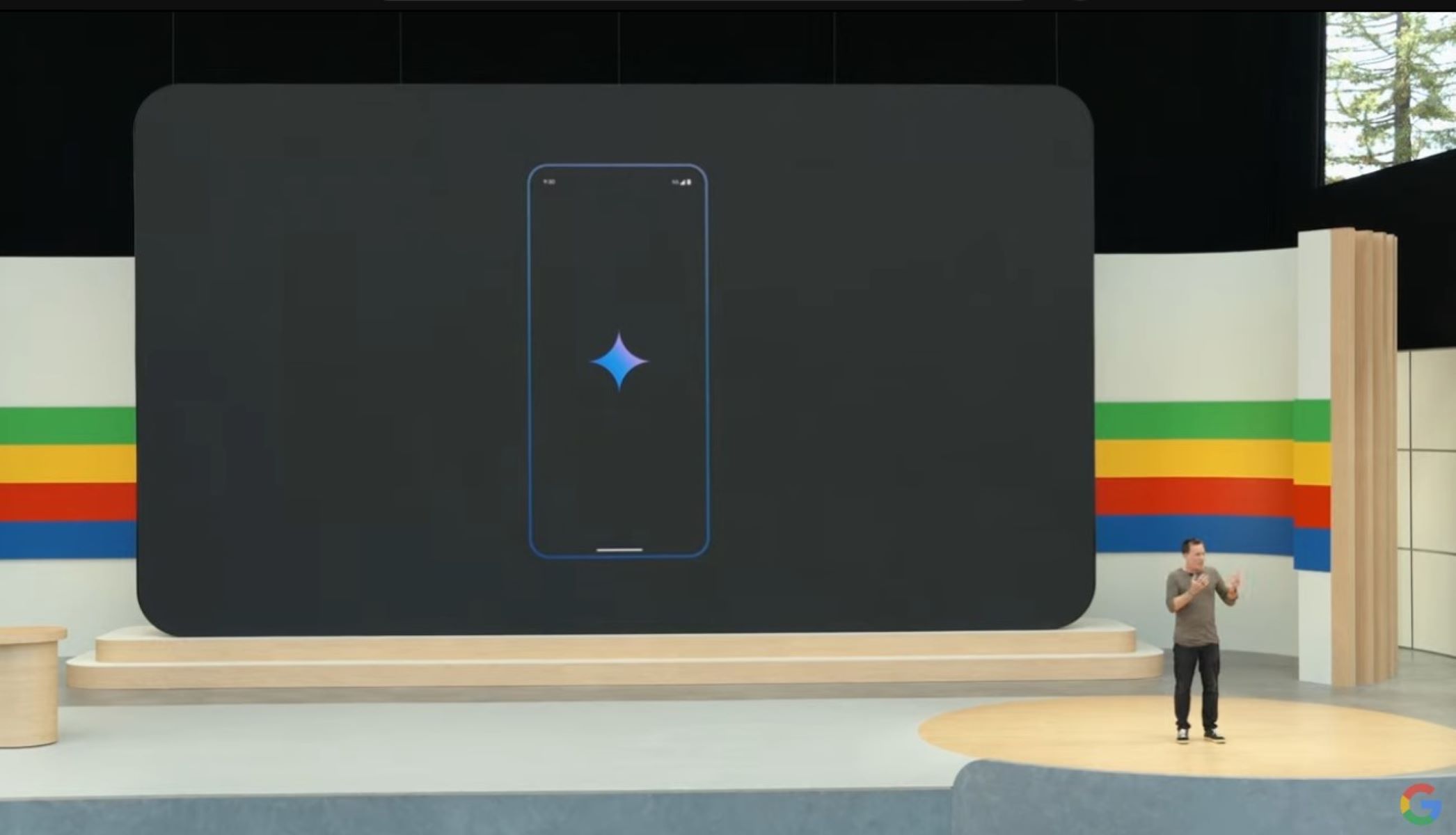 A screenshot of a man standing on stage at Google I/O with an image of the Google Gemini app on a Pixel behind him.