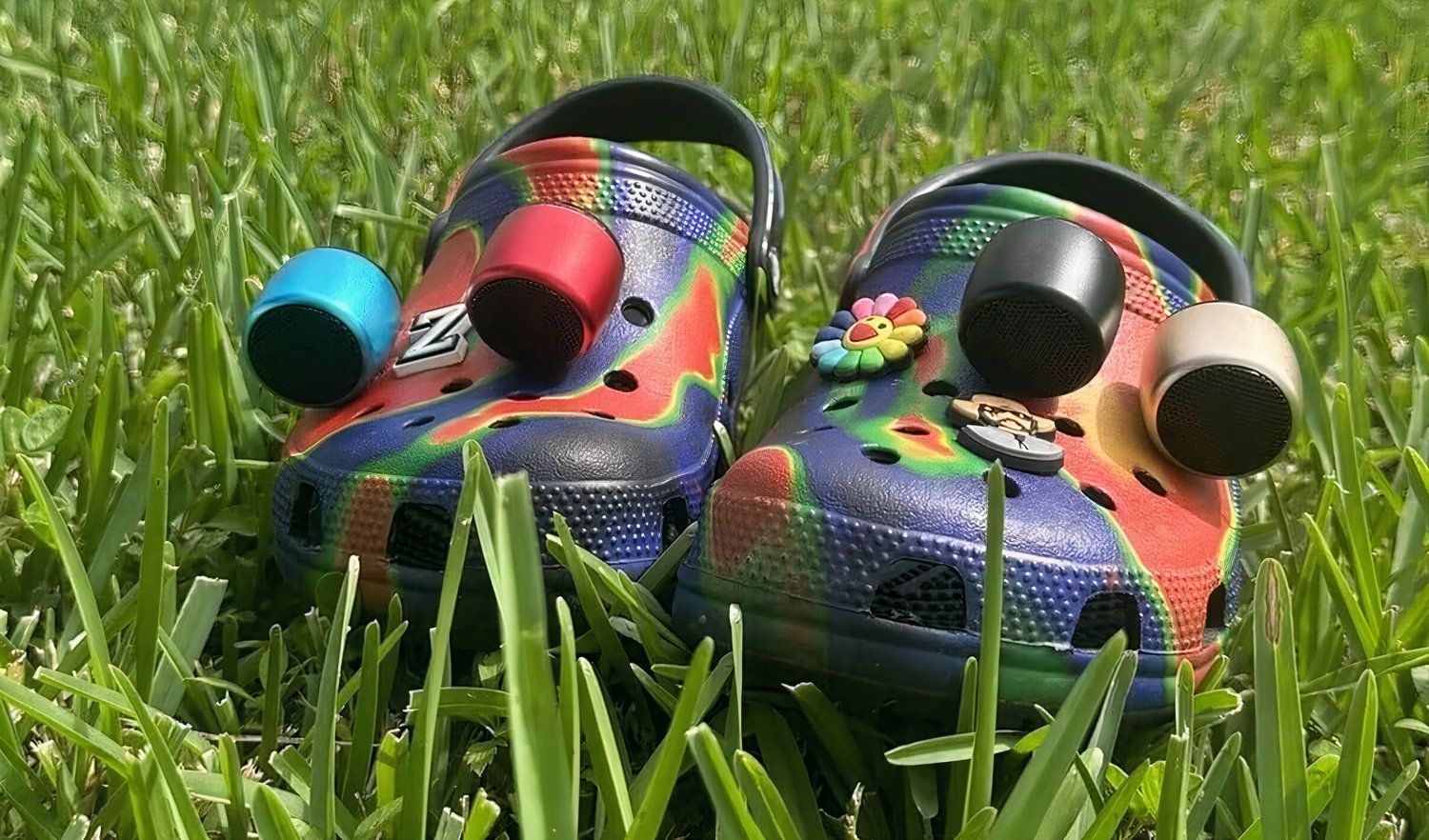 Tie-dye colored Crocs sitting in green grass with Croc charms and tiny bluetooth speakers